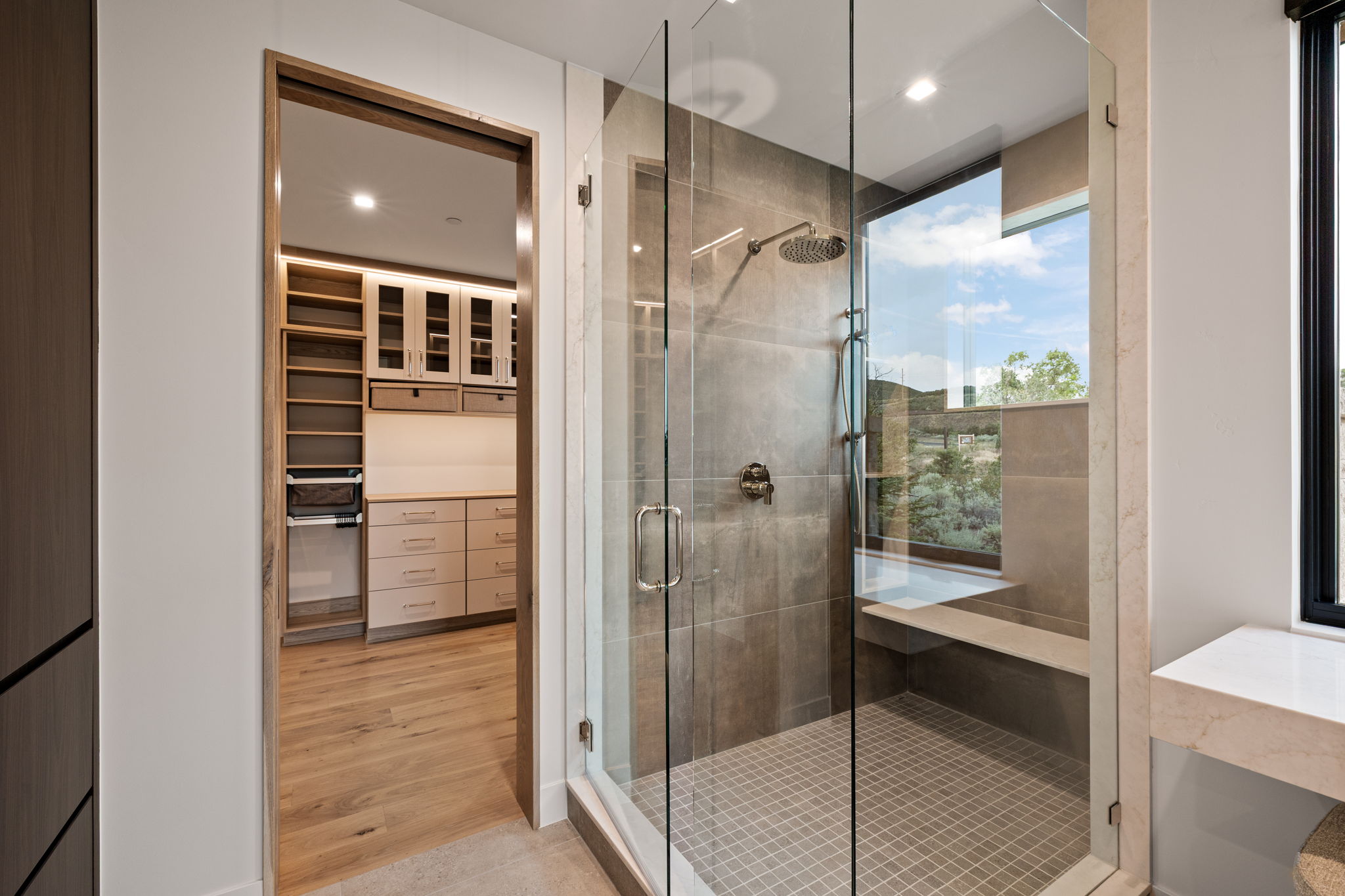 Glass-enclosed shower featuring large format wall tile, rain shower head, built-in bench, and exterior window