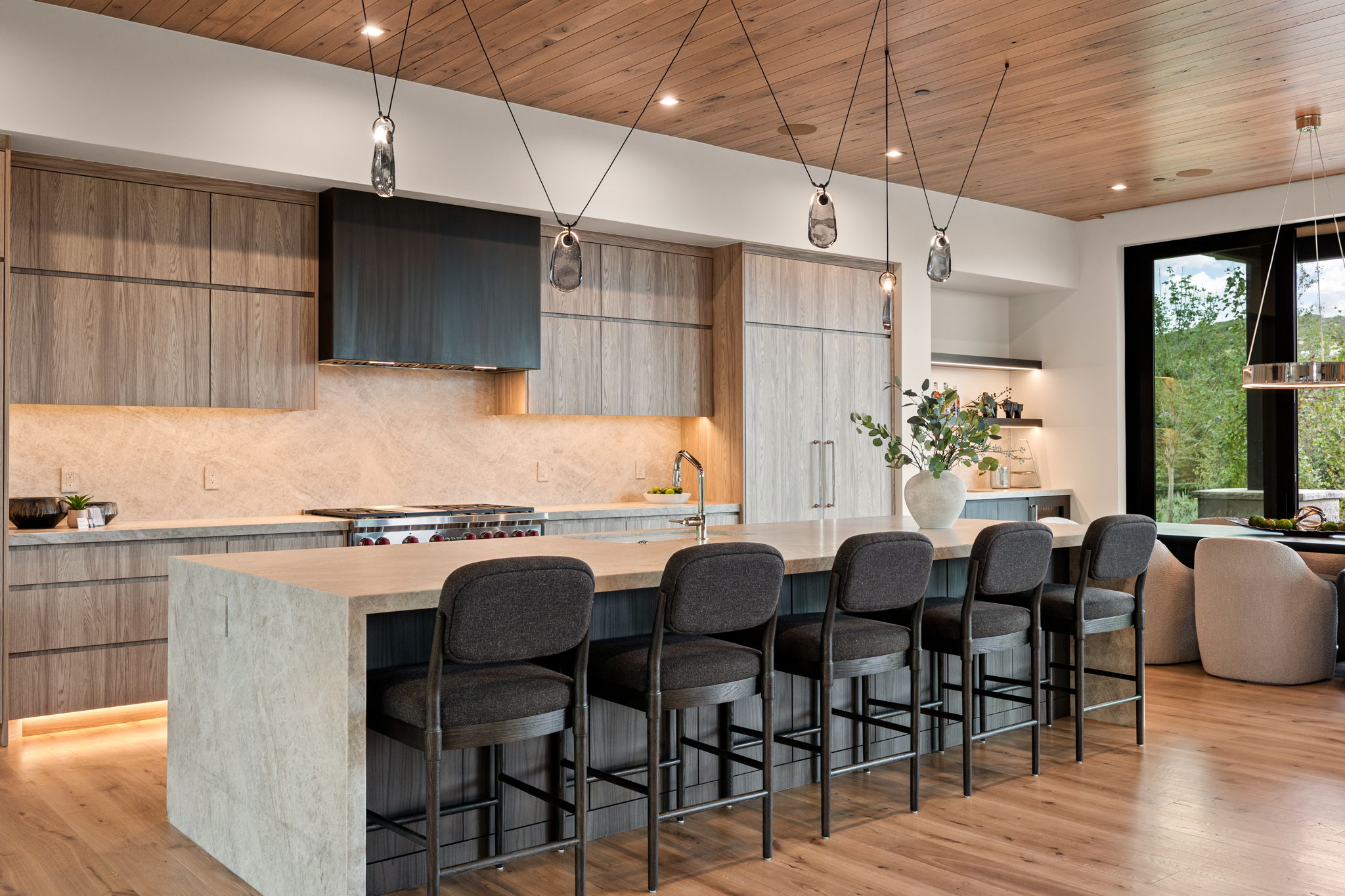 Contemporary kitchen featuring a large waterfall island with seating for five, integrated appliances, wood-finish cabinetry, a gas range with custom hood, and a light wood plank ceiling