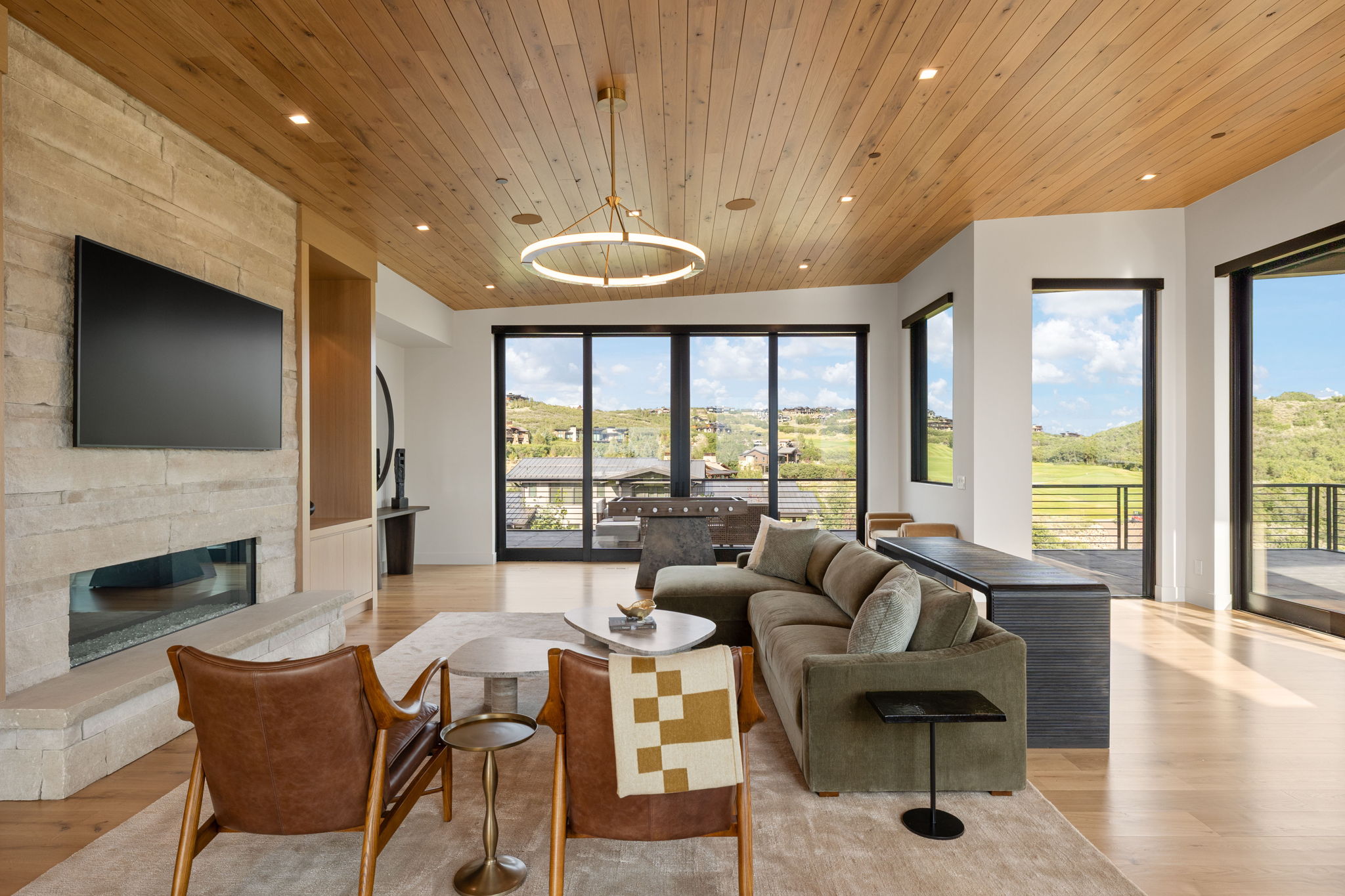 Spacious living area featuring a stone-clad fireplace with an integrated linear firebox, wood-finish flooring, a wood-paneled ceiling, recessed lighting, and expansive glass doors opening to an outdoor deck