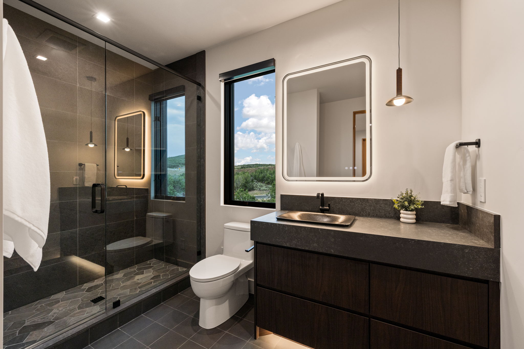 Contemporary bathroom featuring a dark wood-finish floating vanity with a vessel sink, an illuminated framed mirror, and a glass-enclosed shower with dark tile and hexagonal floor tiling