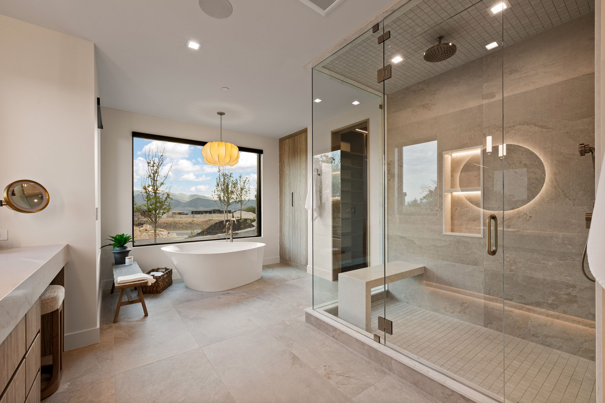 Spacious bathroom featuring a freestanding soaking tub under a large picture window, a large walk-in shower with a built-in bench, a custom stone-finish vanity, and integrated recessed lighting