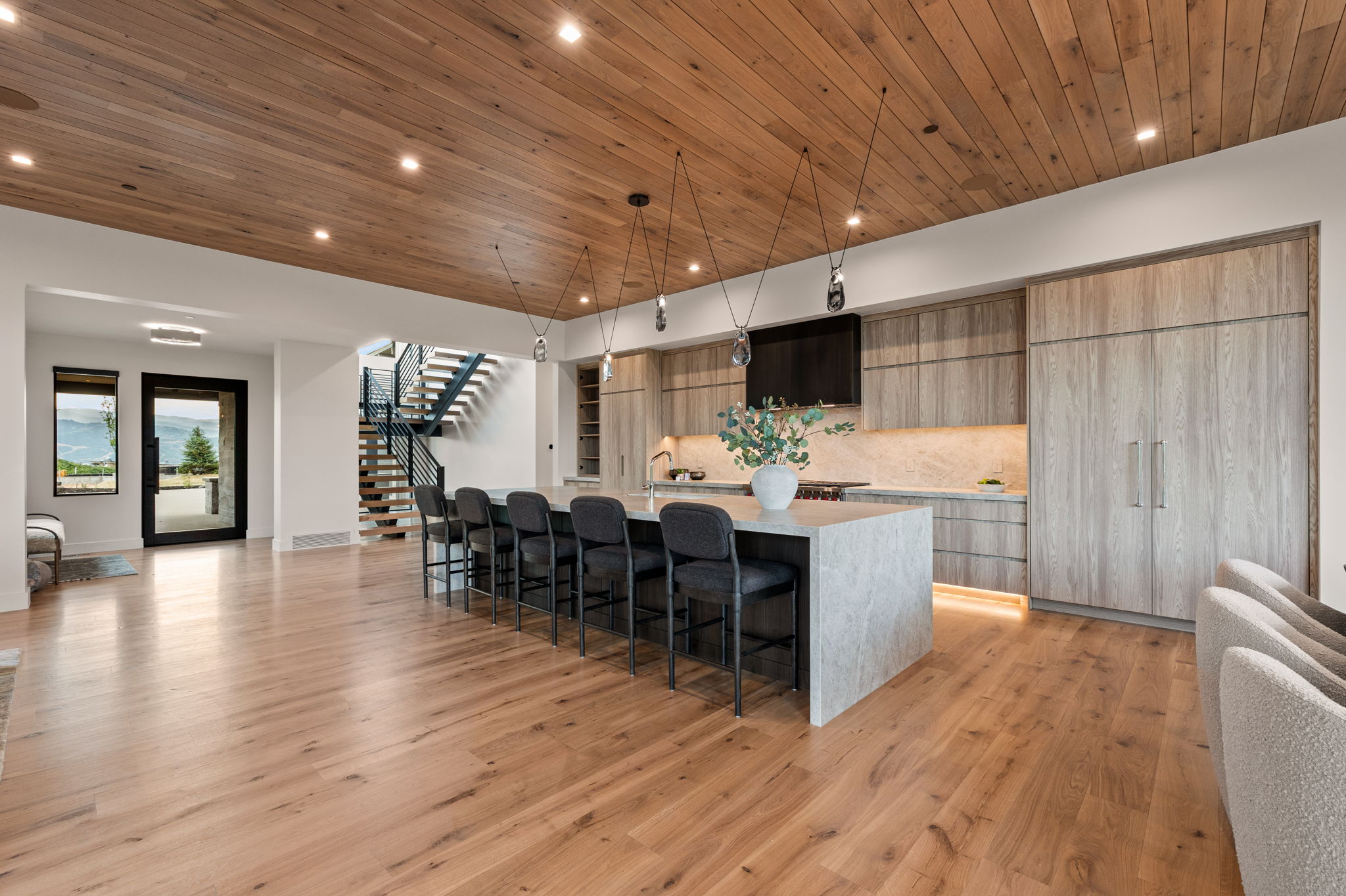 Open-concept living area featuring a kitchen with a large island and integrated appliances, wood-finish flooring, a wood-paneled ceiling with recessed lighting, and a contemporary staircase