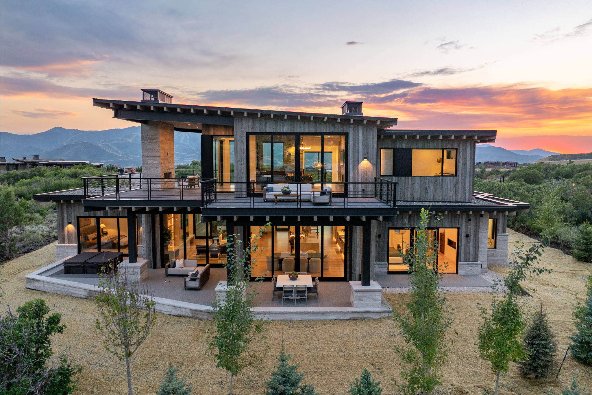 Multi-level contemporary residence featuring extensive glass doors and windows, multiple outdoor patios and balconies, a distinctive sloped roofline, exposed stone accents, and wood-finish siding
