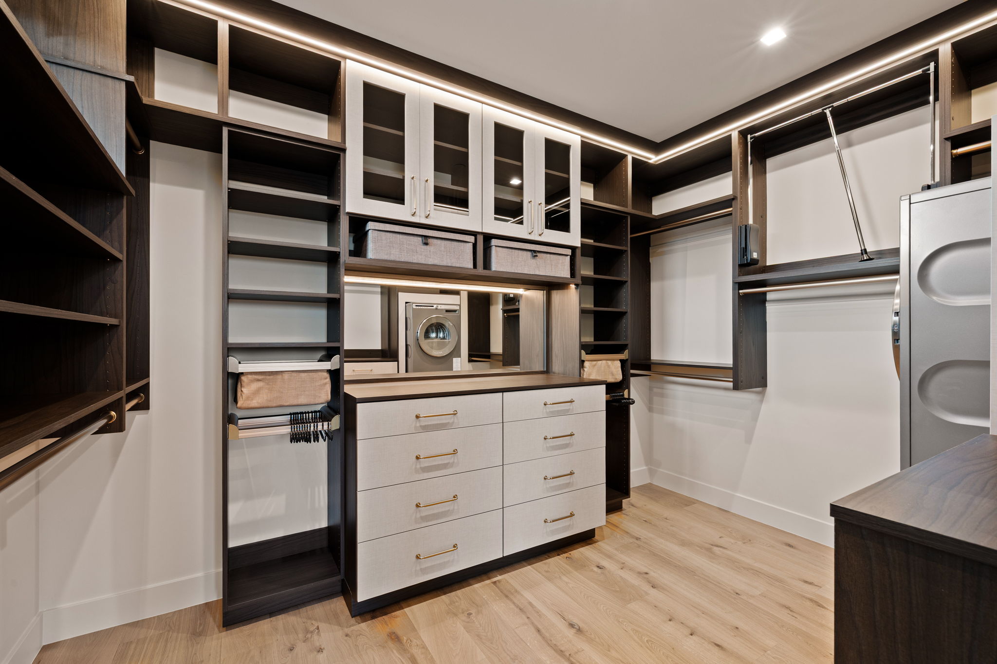 Custom walk-in closet featuring built-in cabinetry, integrated lighting, wood-finish flooring, a central dresser, and glass-front upper cabinets