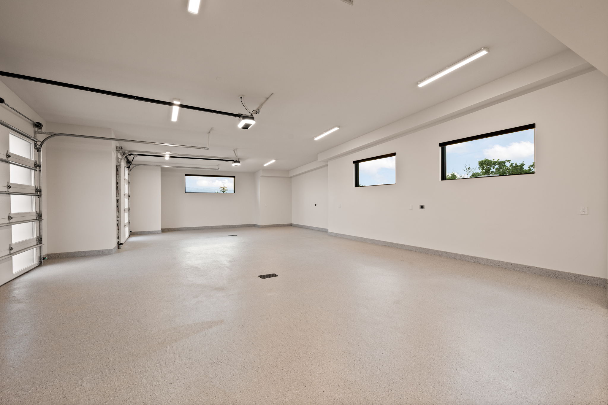 Spacious garage with light-colored epoxy flooring, three high-set windows, multiple linear ceiling lights, and two glass-paneled automatic garage doors