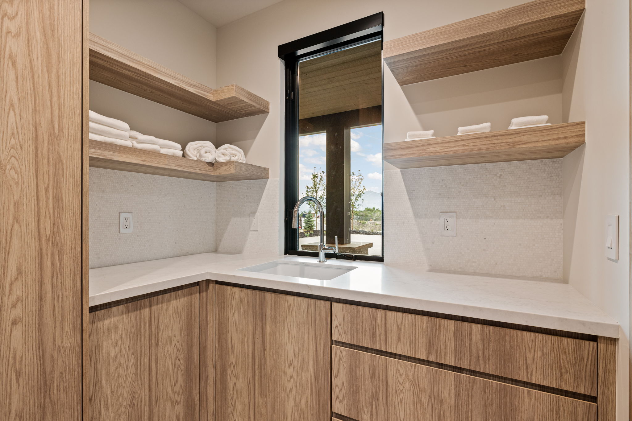 Laundry area with a corner sink, light wood-finish cabinetry, white countertop, and open shelving