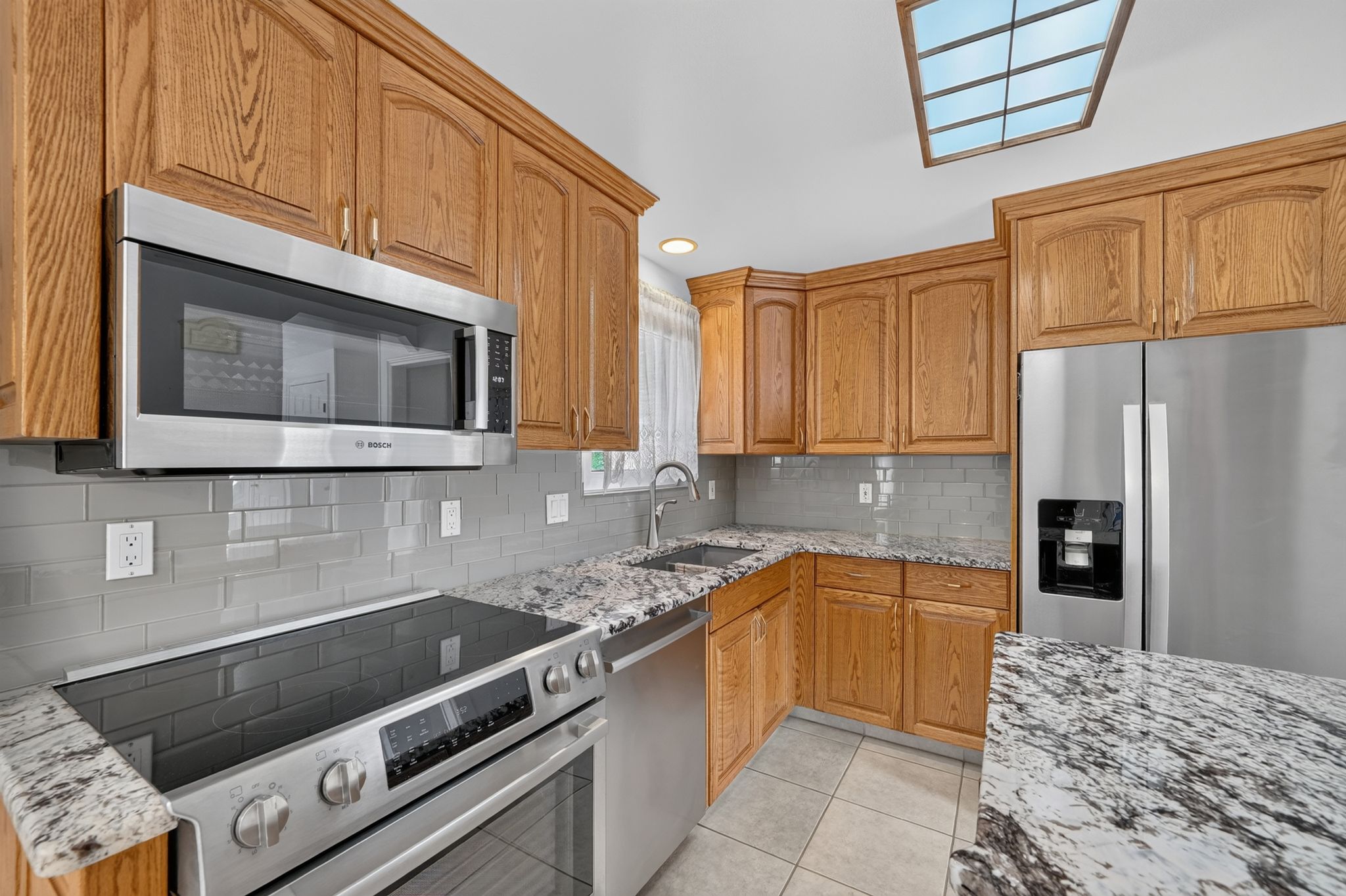 Kitchen featuring wood cabinetry, stainless steel appliances, grey subway tile backsplash, light-toned granite countertops, and ceramic tile flooring