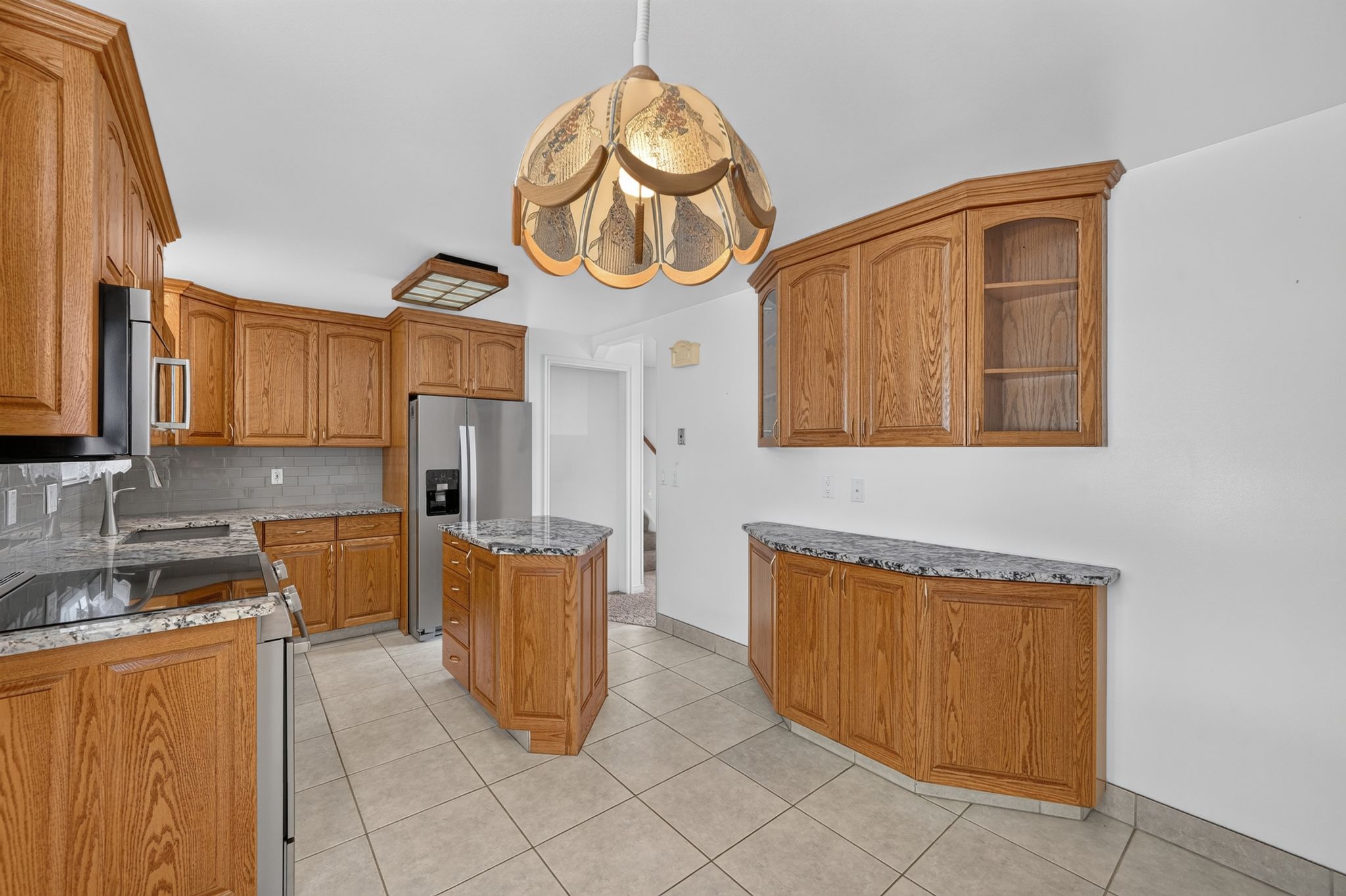 Kitchen featuring wood cabinetry, light-toned tiled flooring, a central island with a stone-finish countertop, stainless steel appliances, and a tiled backsplash