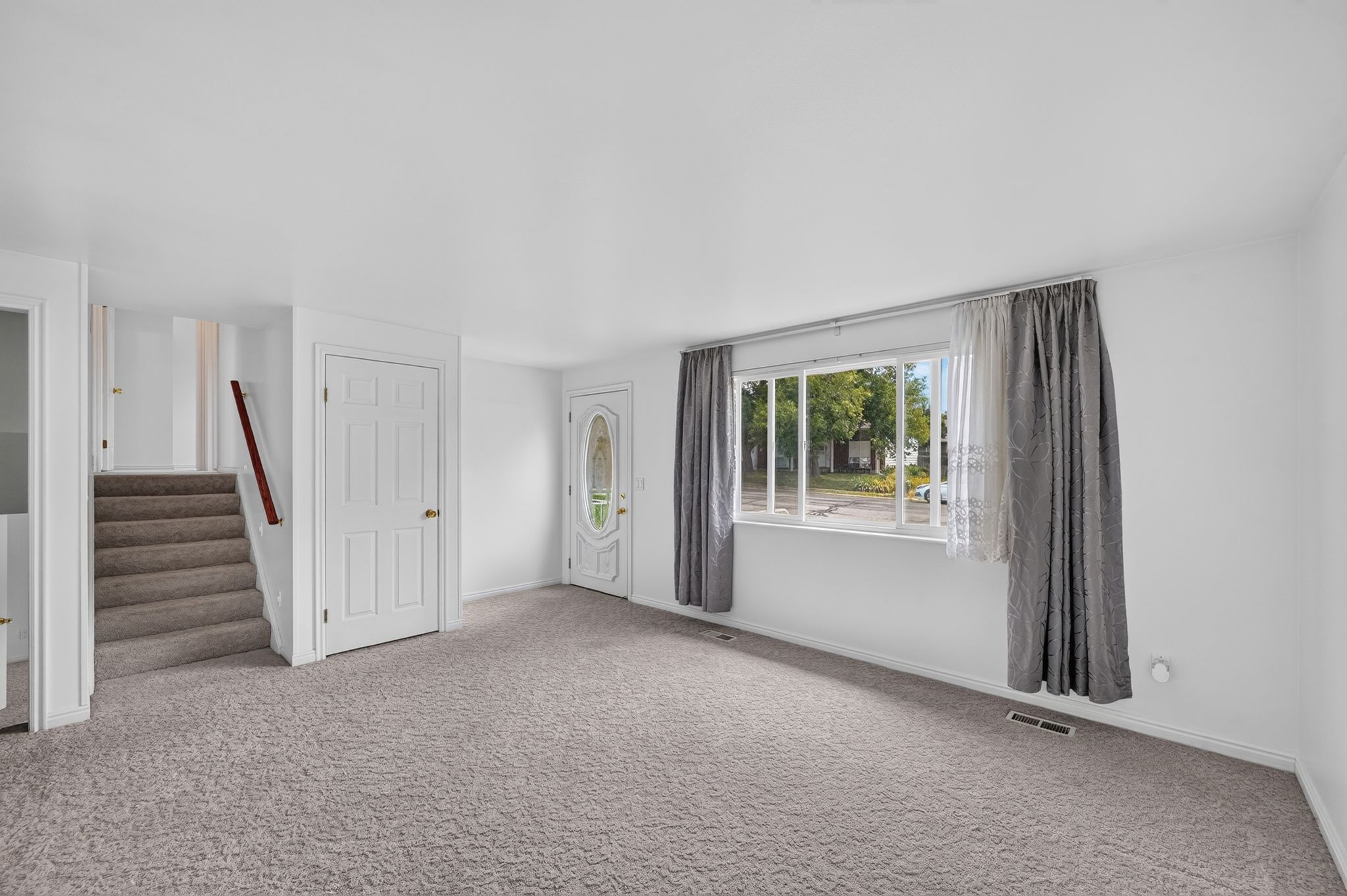 Spacious room featuring neutral carpeting, a large multipane window, a paneled door, a front entry door with decorative glass, and a carpeted staircase with a wood handrail