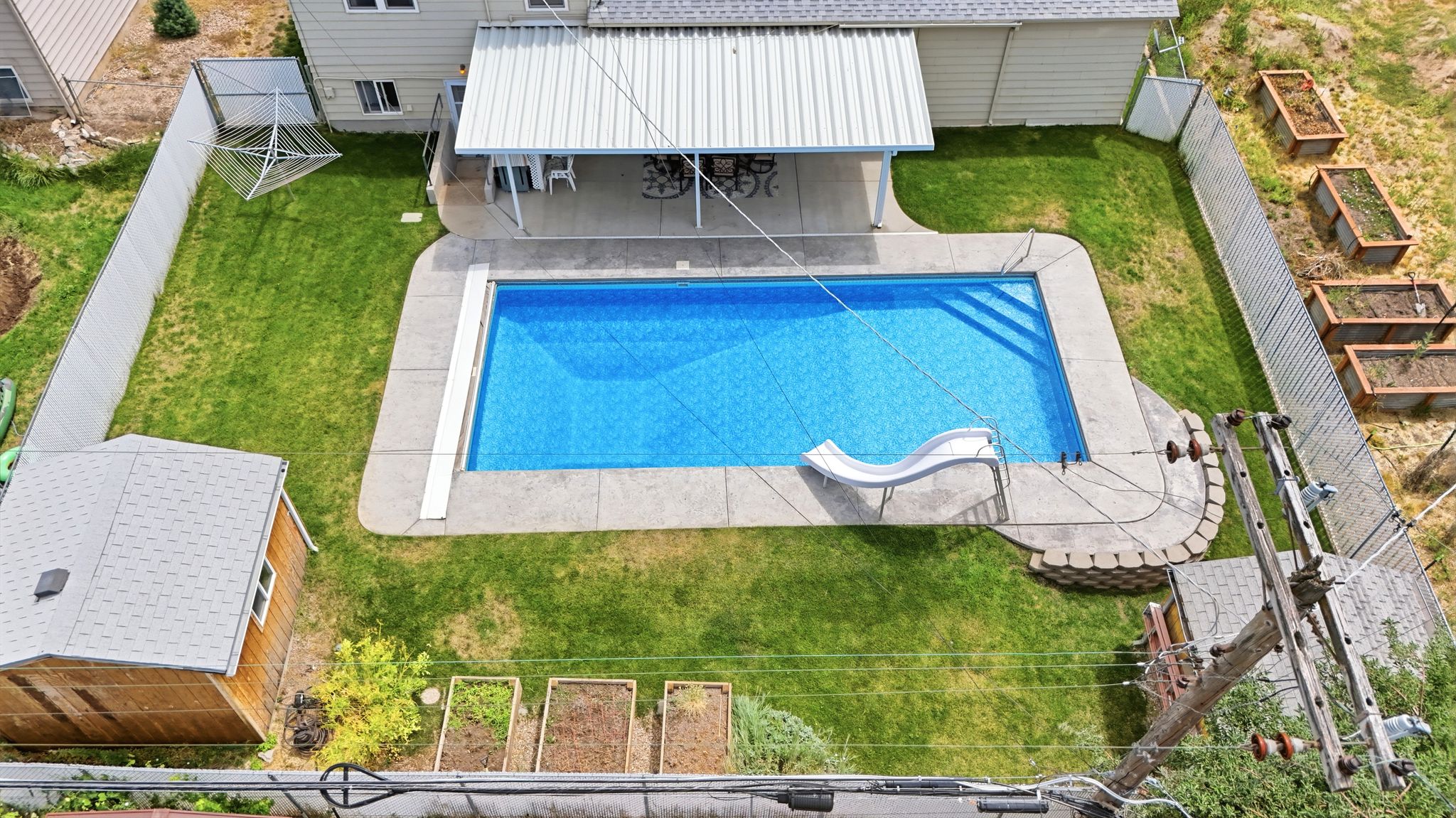 Expansive backyard featuring a rectangular swimming pool with an integrated slide, a concrete patio area with an overhead metal awning, and a separate wood-sided shed with a shingled roof
