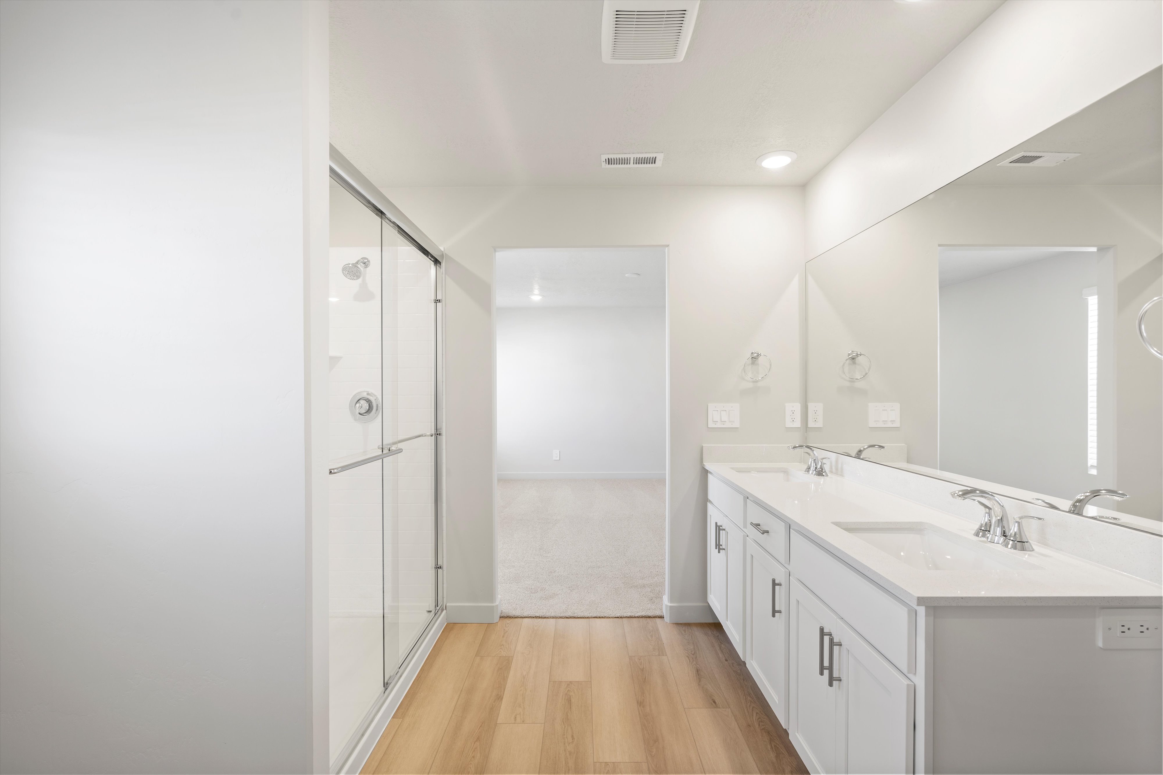 Spacious bathroom featuring a dual vanity with white cabinetry and an integrated countertop, a large wall-mounted mirror, a glass-enclosed shower, and wood-finish flooring