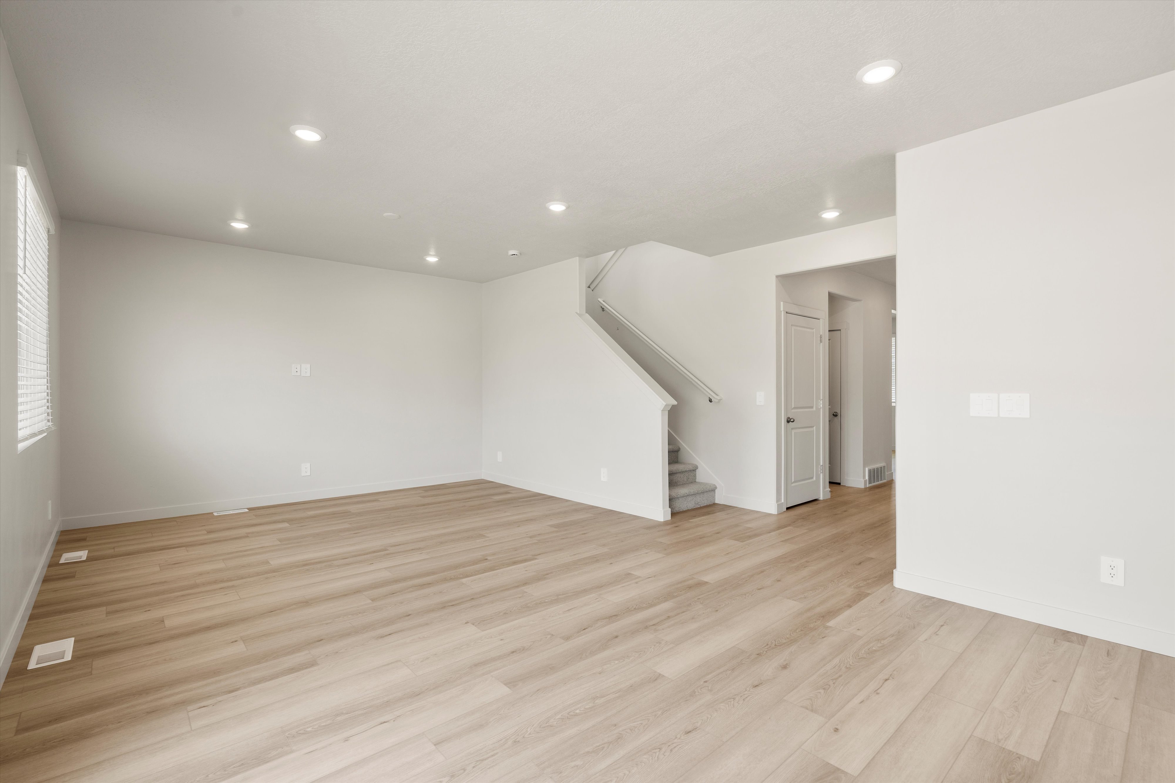Spacious open-concept room featuring light wood-finish flooring, recessed lighting, white walls, and a staircase with carpeted treads and a handrail