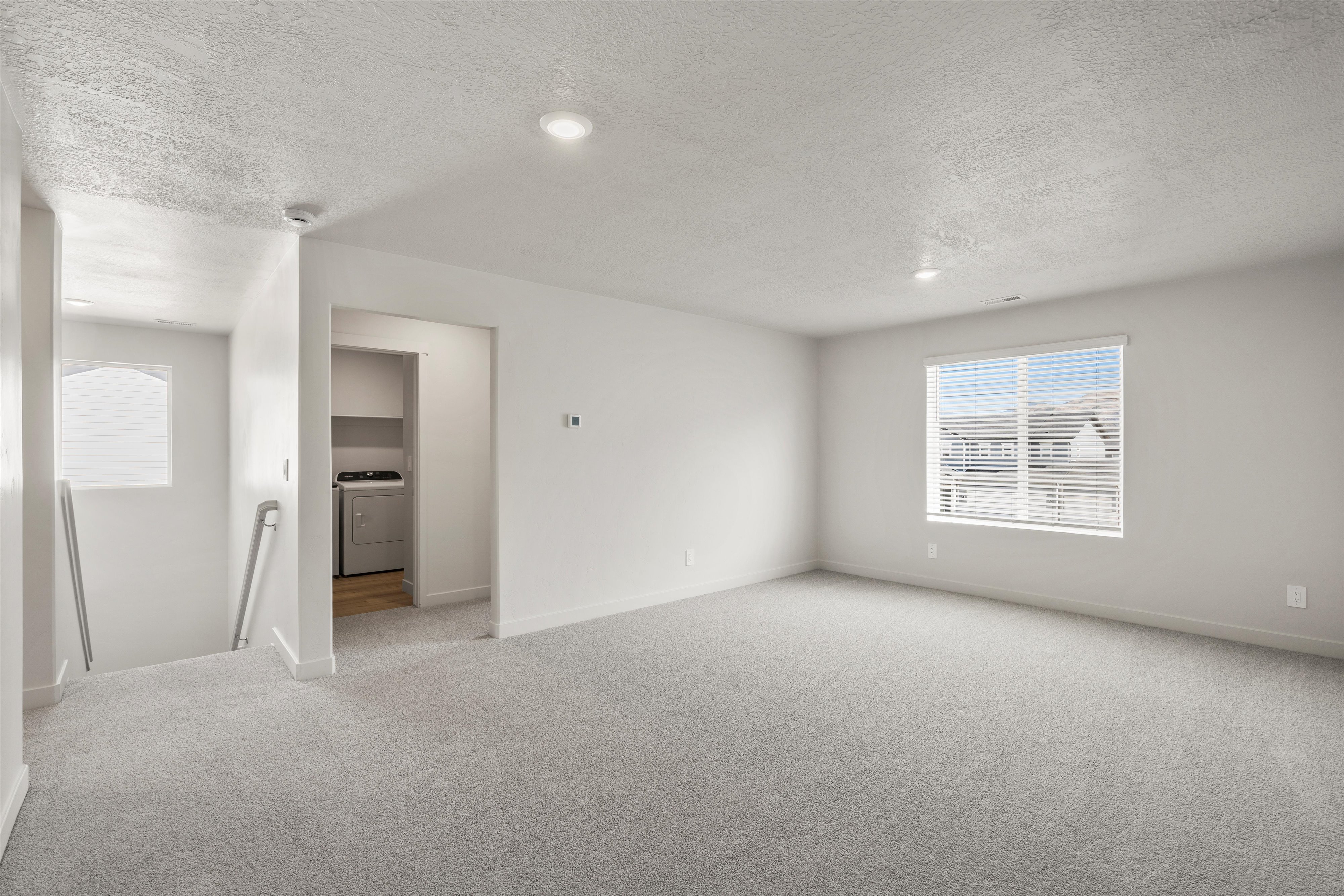 Spacious room featuring light gray carpeting, recessed lighting, and a large window with horizontal blinds