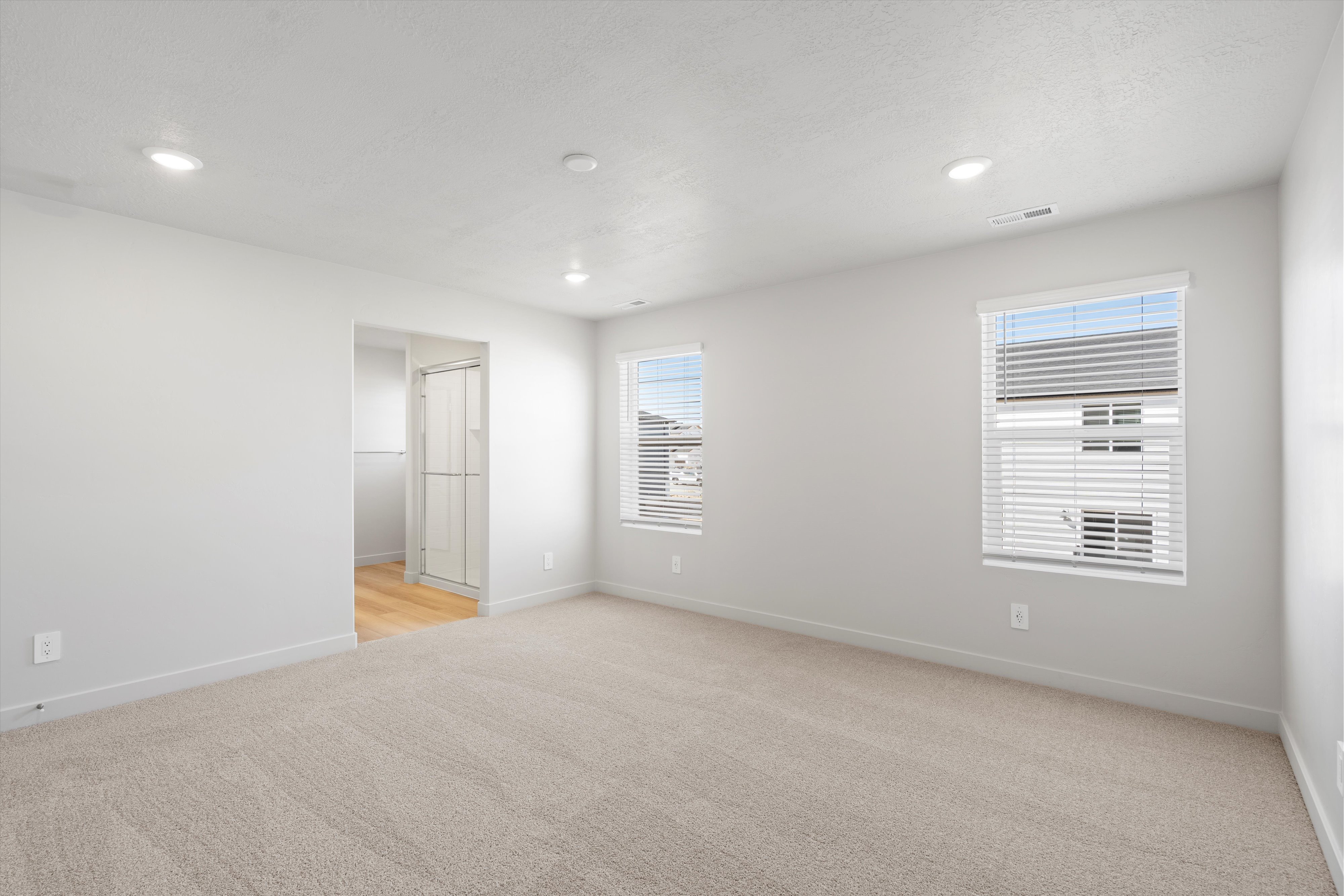 Carpeted room featuring recessed lighting, two windows with blinds, and an entryway to a space with wood-finish flooring and a glass-enclosed shower