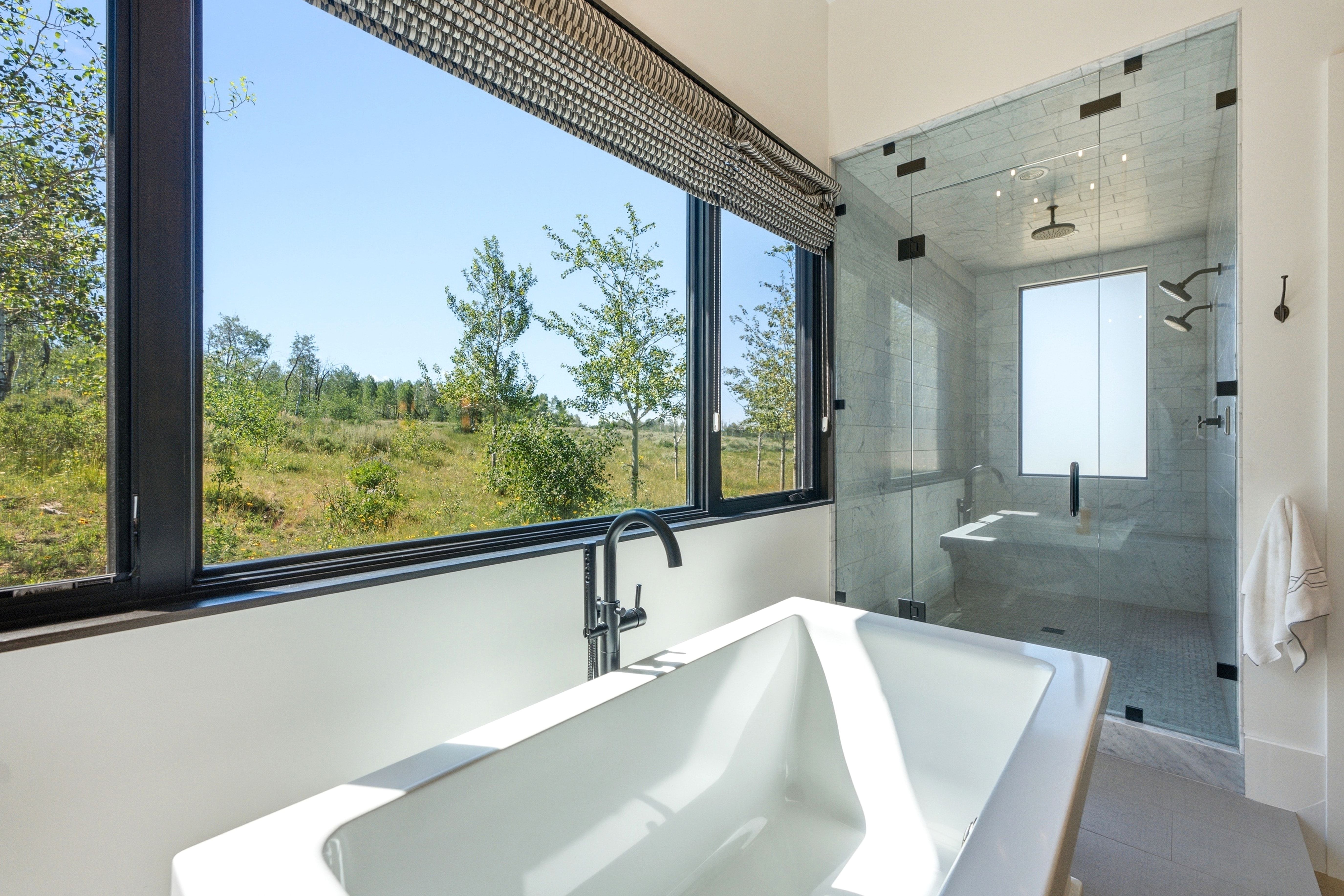 Spa-inspired bathroom featuring a freestanding soaking tub with a modern faucet, a spacious glass-enclosed shower with multiple showerheads and an integrated bench, and a large window with roller shades overlooking natural landscape