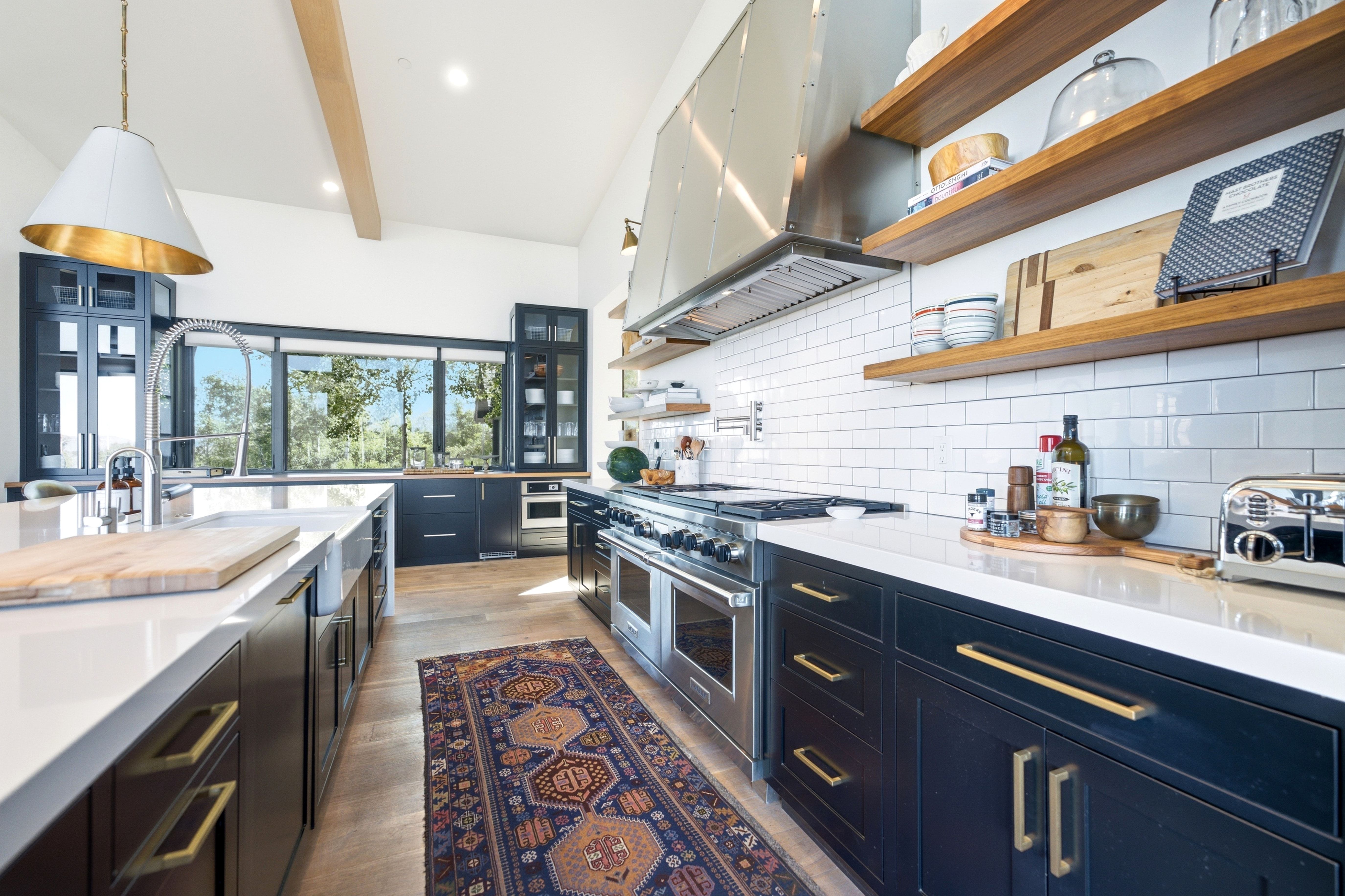 Kitchen featuring a large central island with a farmhouse sink, stainless steel appliances, dark cabinetry, open wood shelving, and extensive window-line