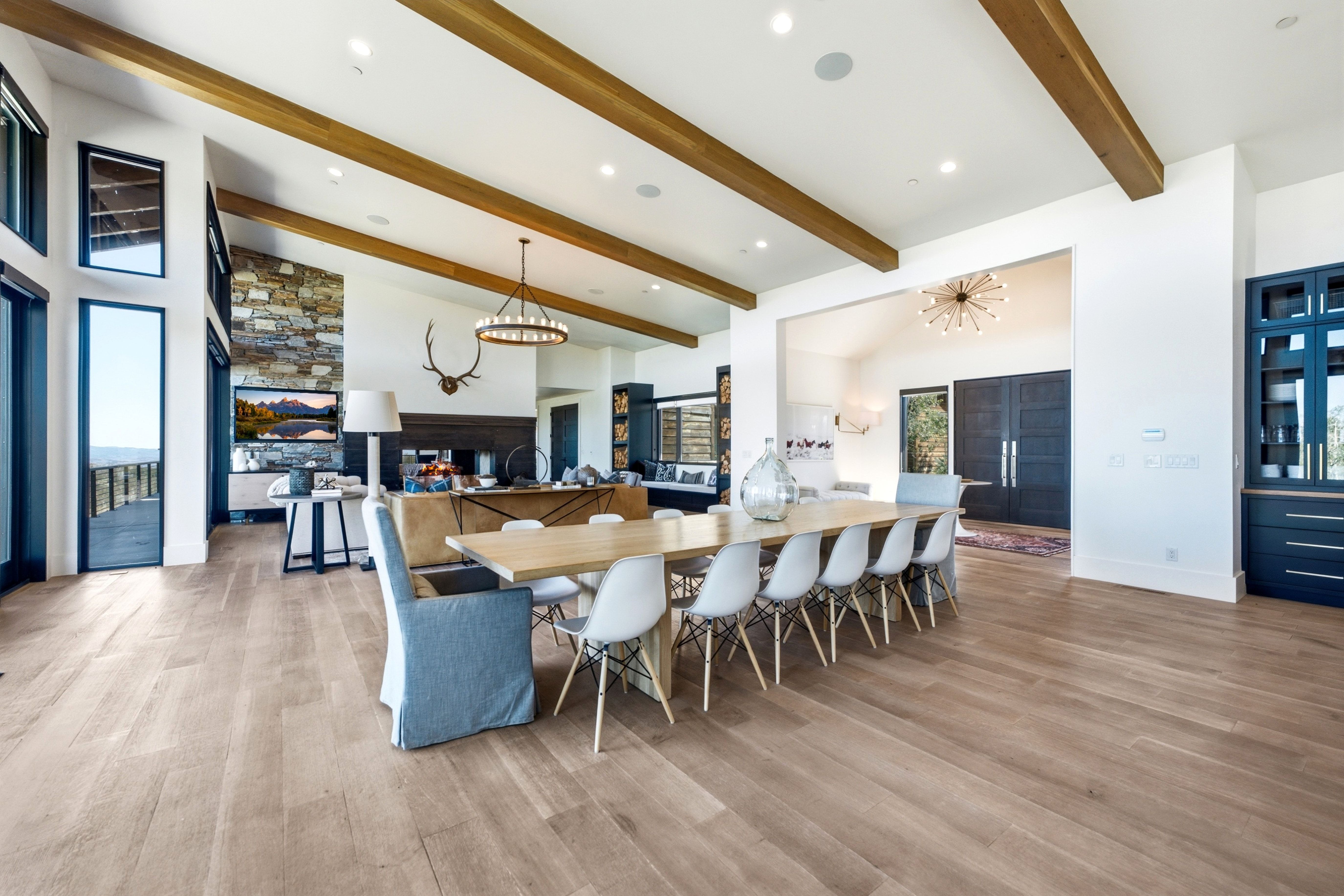 Spacious interior featuring wood-finish flooring, vaulted ceilings with exposed wood beams, a stone accent wall, large floor-to-ceiling windows, and a built-in dark wood cabinetry with glass fronts