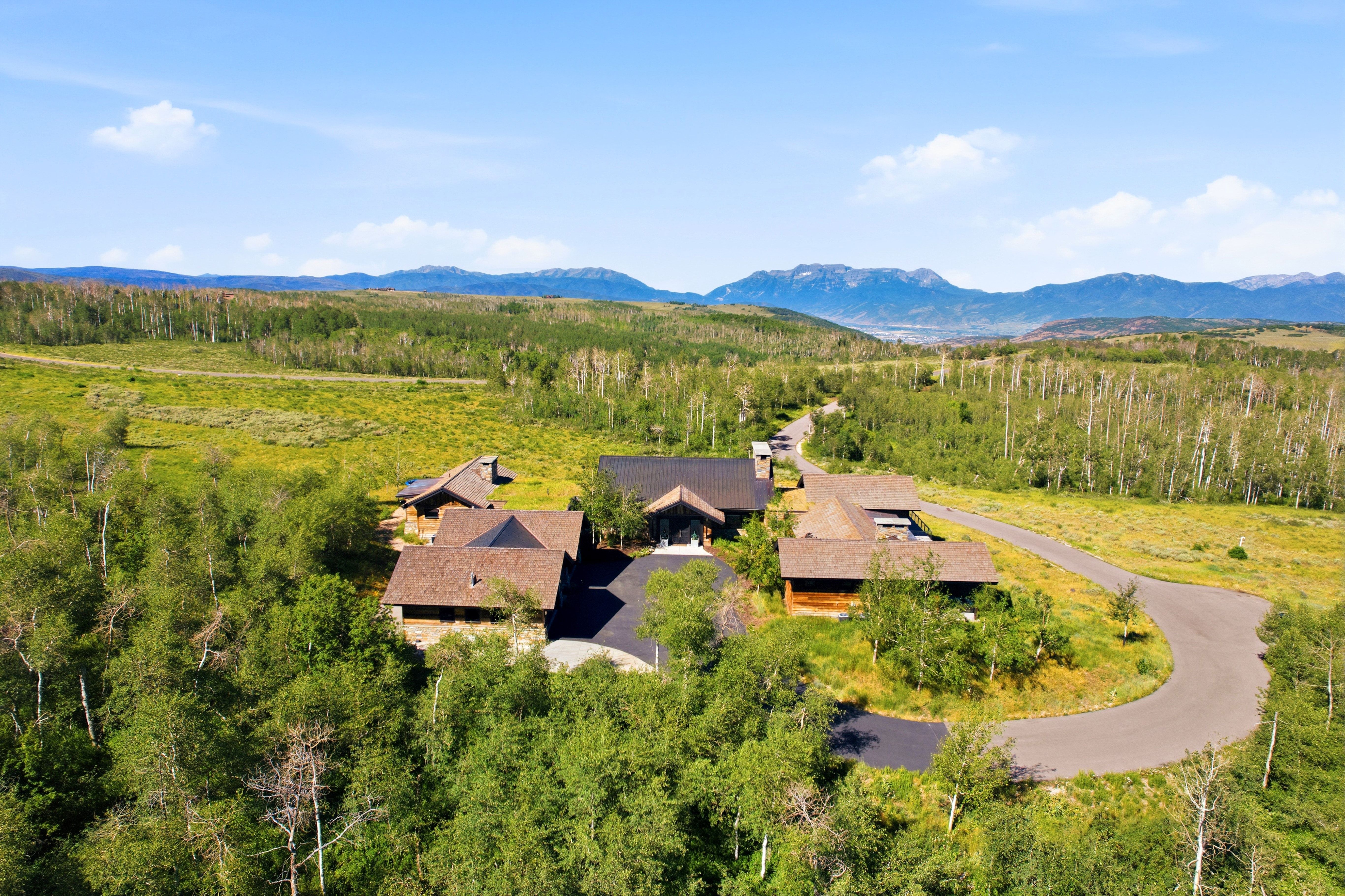 Expansive property featuring multiple structures with dark roofs and natural wood siding, surrounded by mature trees and rolling green terrain