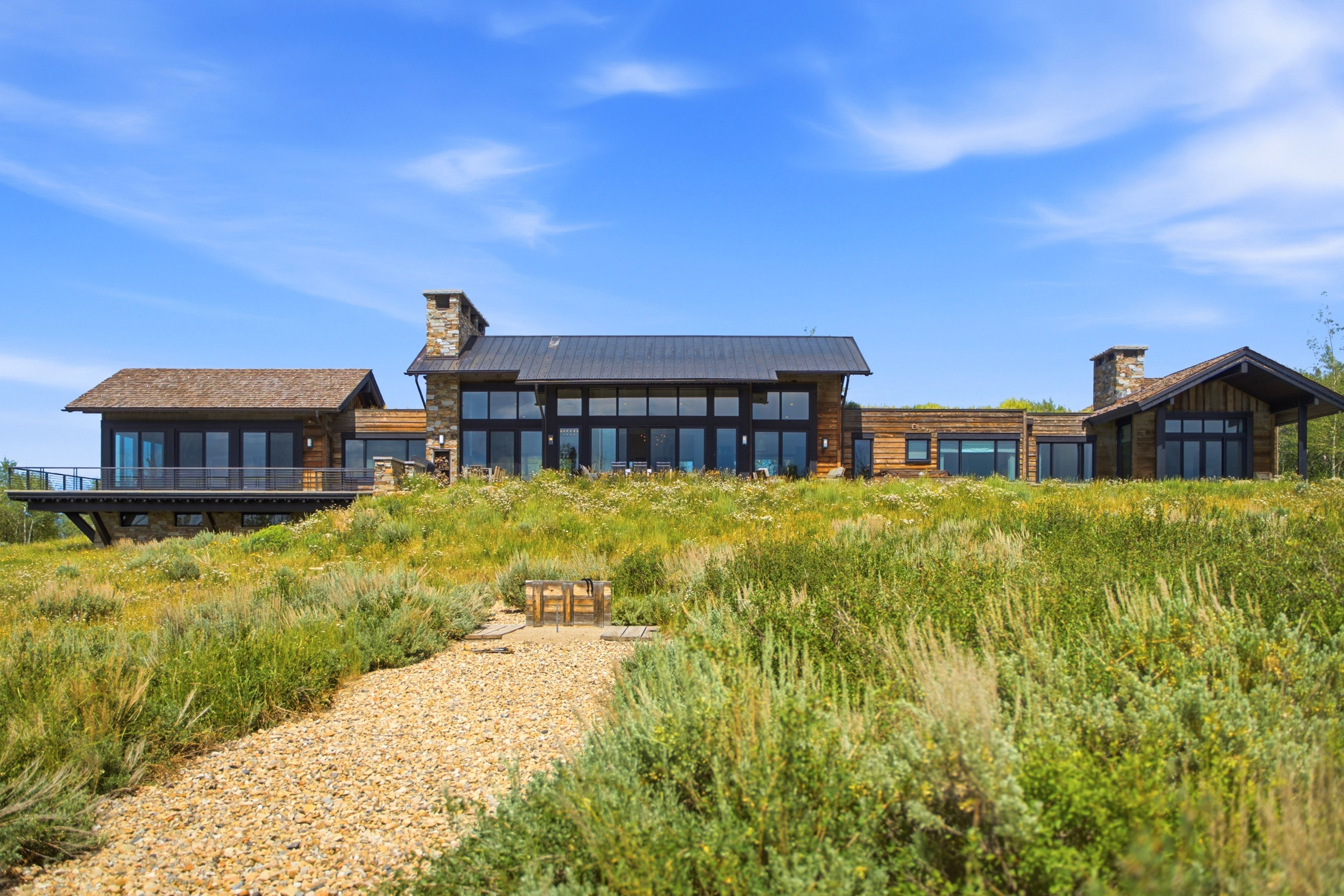 Expansive contemporary residence featuring wood and stone facade, large windows, dark metal roof, and a gravel path