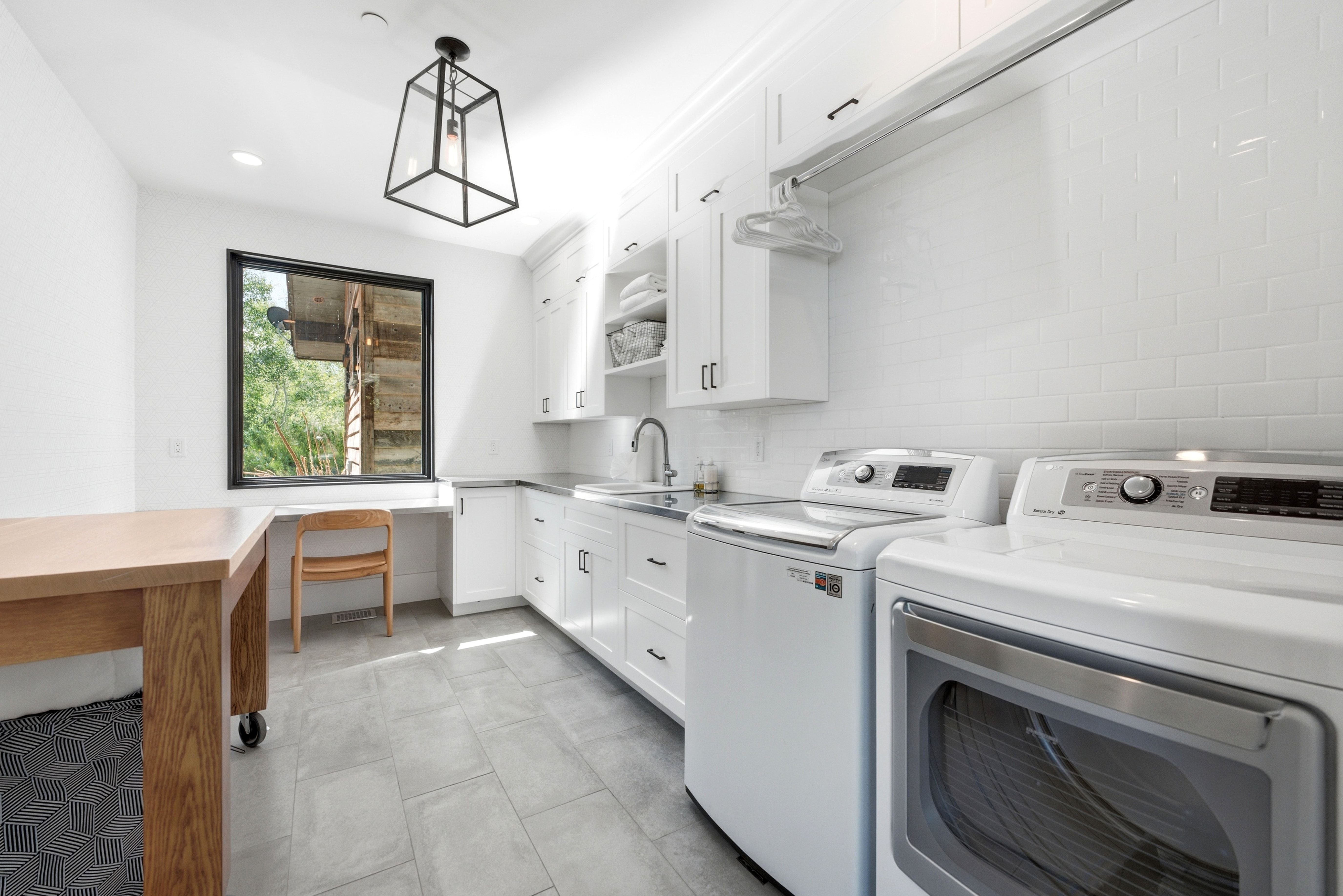 Bright laundry room featuring a large window, white shaker cabinetry, stainless steel sink, subway tile backsplash, and light gray tile flooring