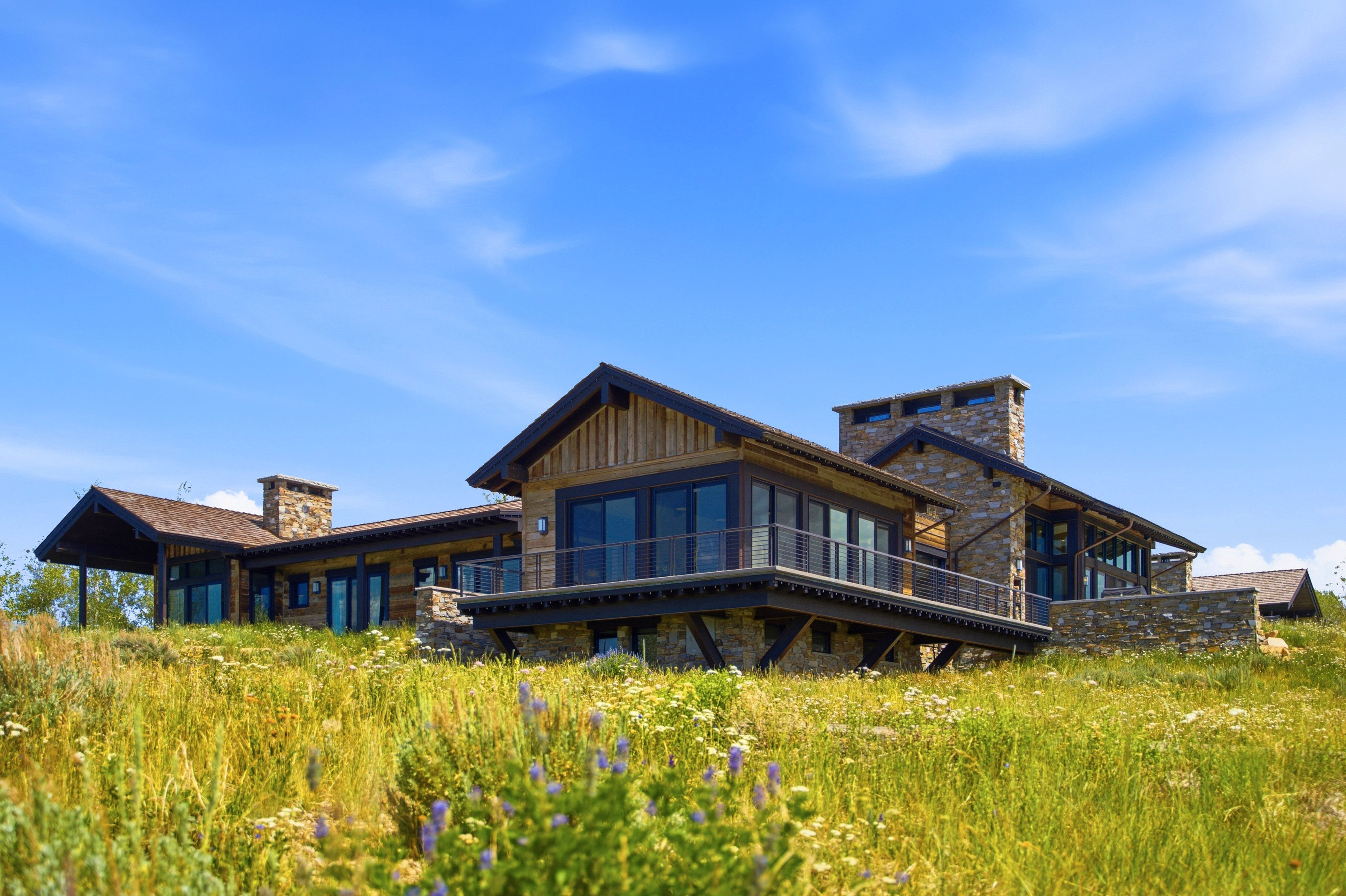 Stone and wood-sided architectural style featuring multiple chimneys, a substantial deck with metal railings, a covered patio, and extensive glass doors and windows
