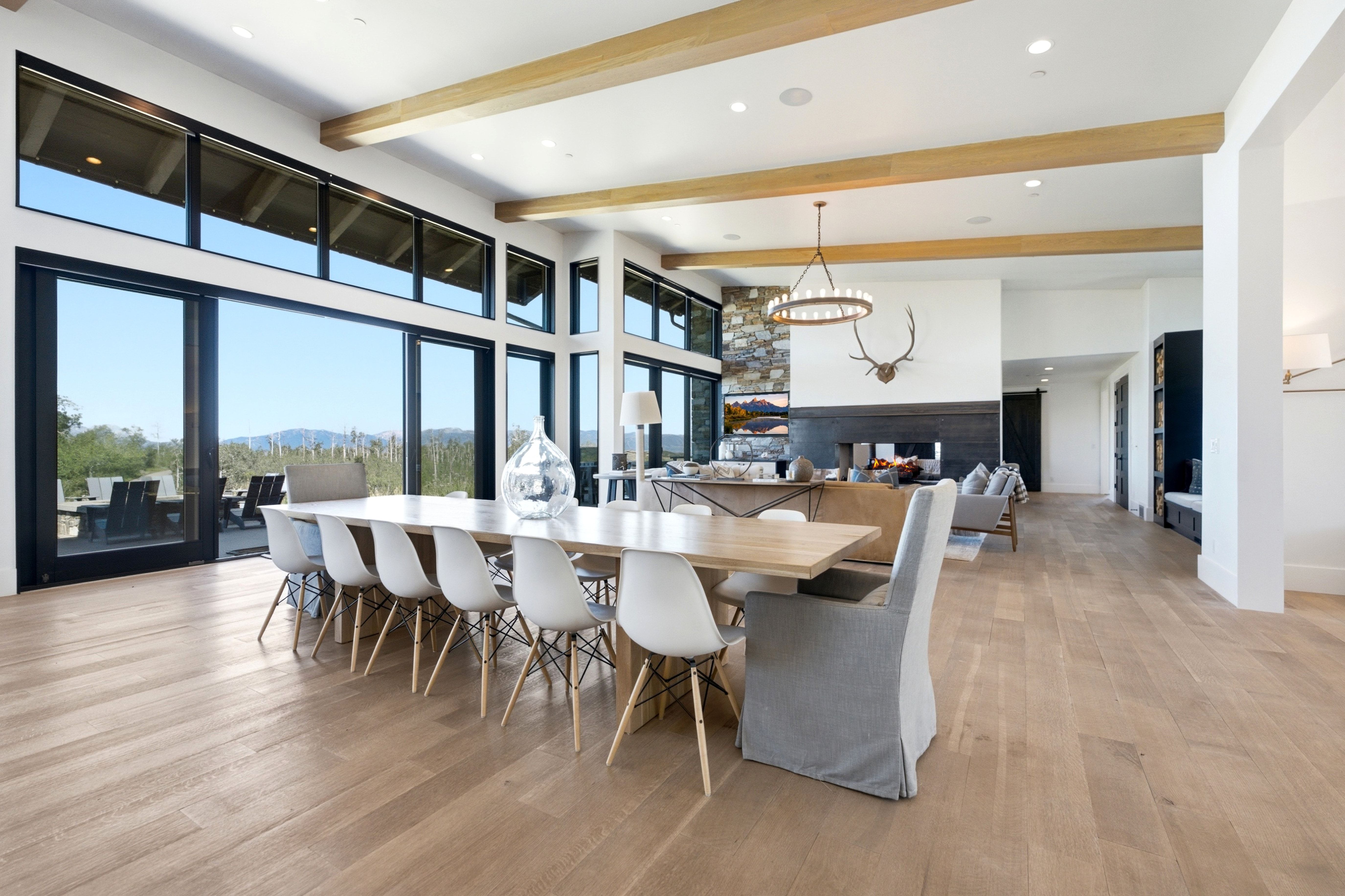 Spacious dining area featuring expansive floor-to-ceiling windows, light wood-finish flooring, exposed ceiling beams, a contemporary chandelier, and a stone accent wall with a linear fireplace