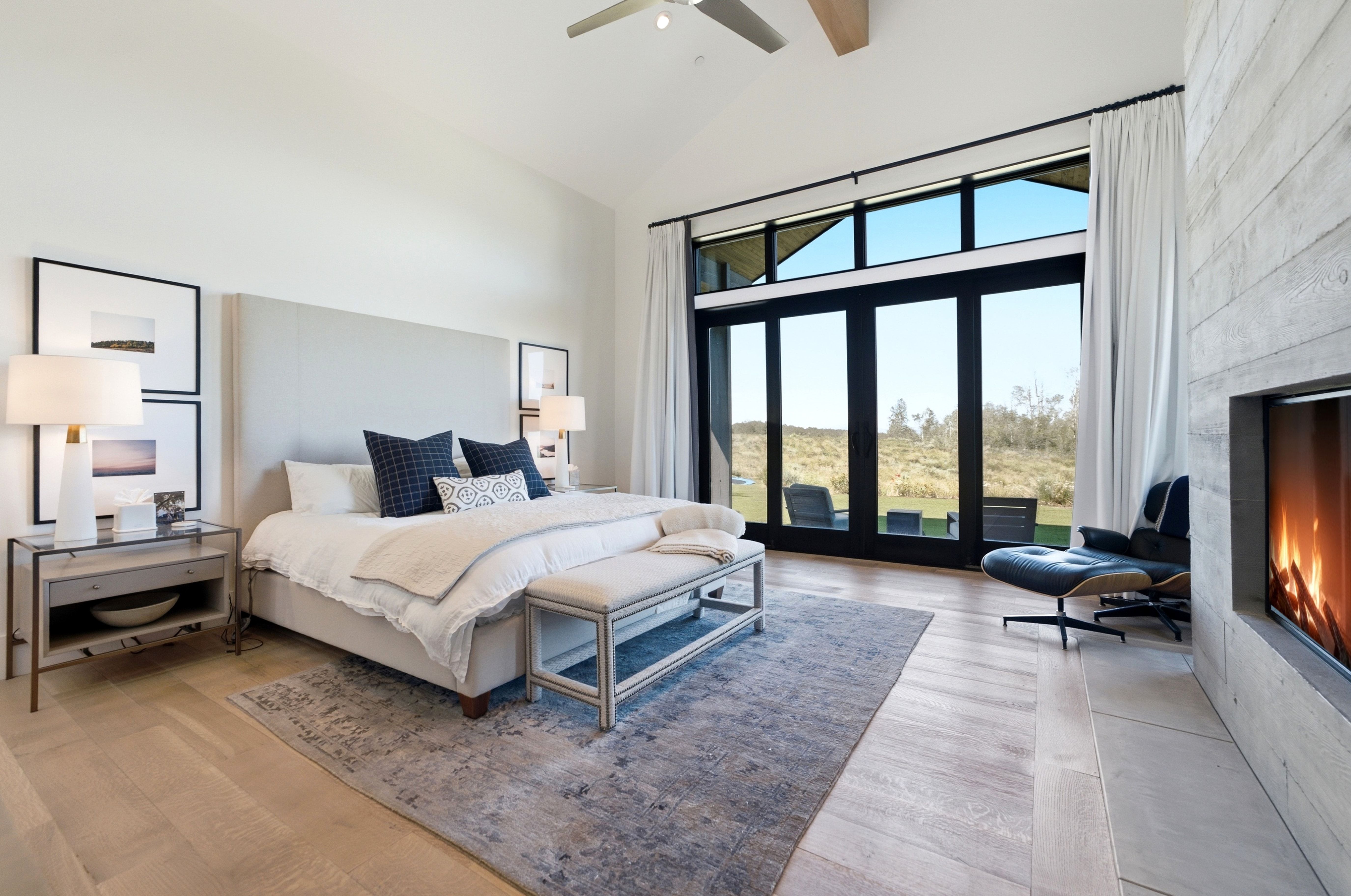 Vaulted ceiling bedroom featuring wood-finish flooring, a full-height upholstered headboard, a built-in fireplace with a textured concrete surround, and expansive black-framed glass doors