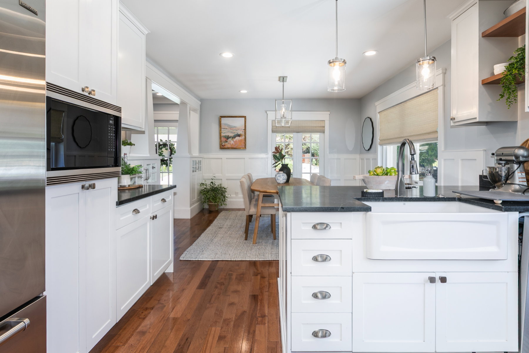 Updated kitchen featuring white shaker cabinetry, an integrated farmhouse sink, dark stone countertops, wood-finish flooring, and stainless steel appliances