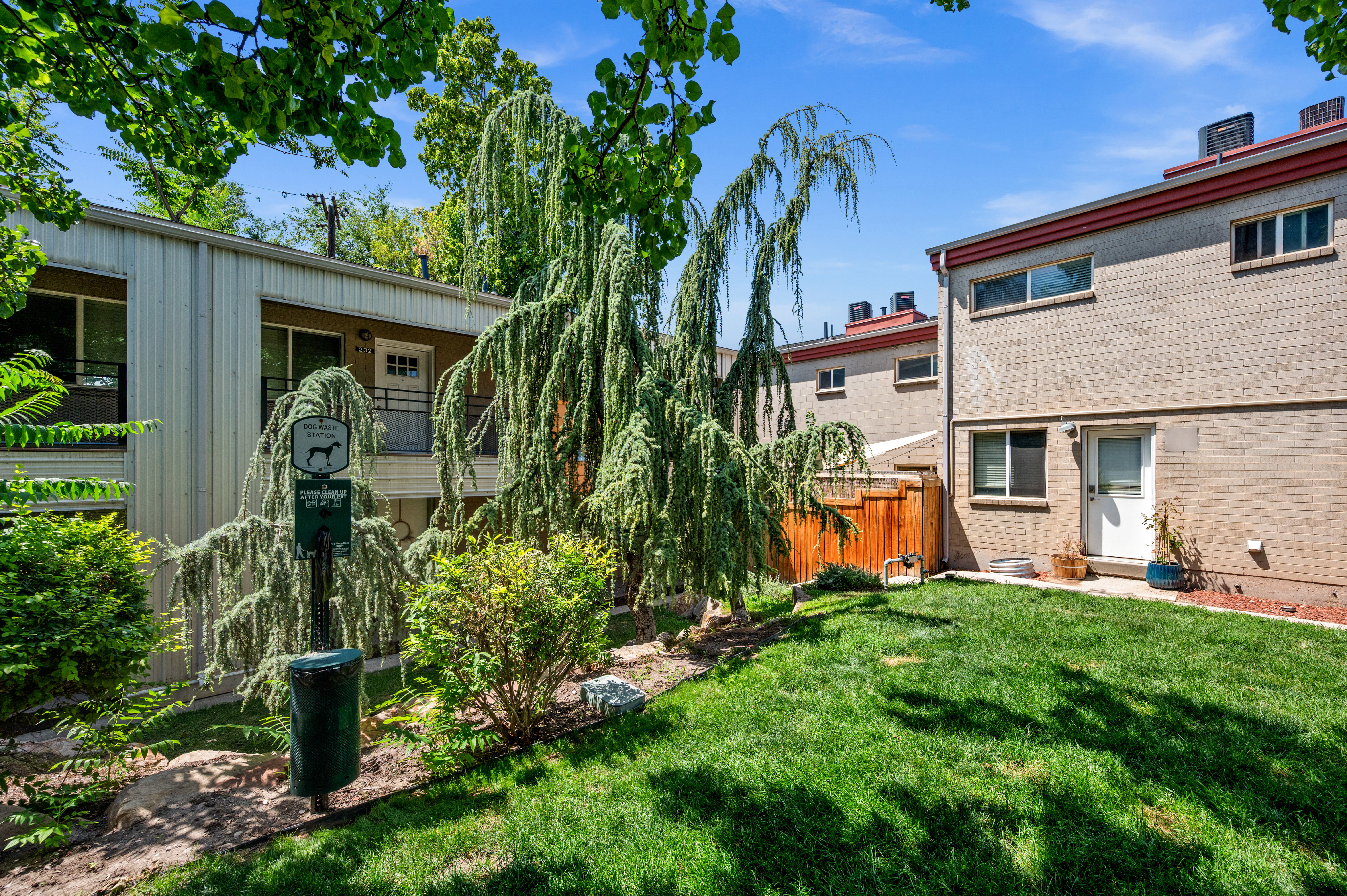 Expansive grassy yard area featuring mature trees and shrubbery, a wood fence, and a brick-faced exterior building