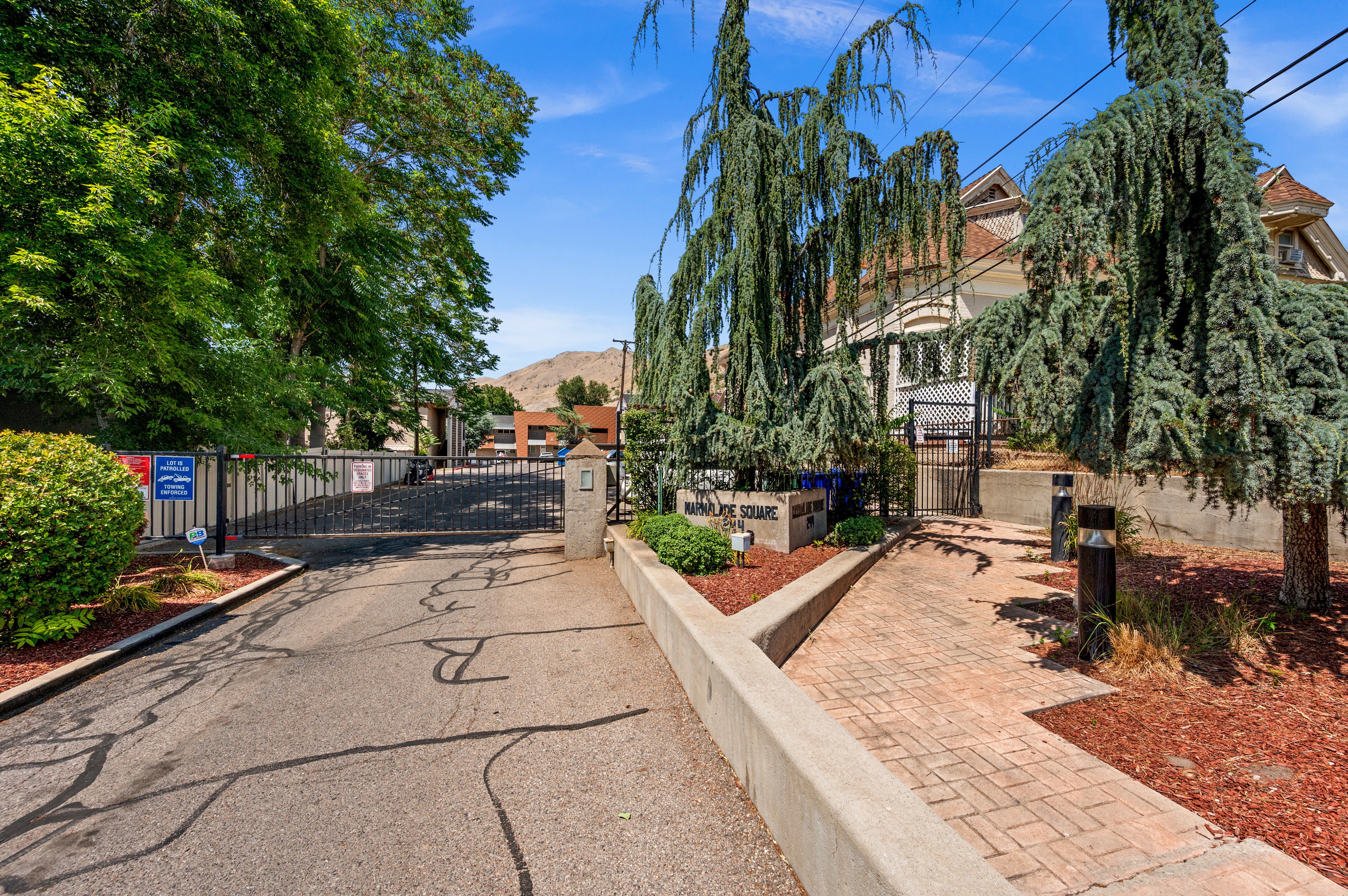 Gated community entrance featuring an asphalt drive, stone pillars, and manicured landscaping with mature trees