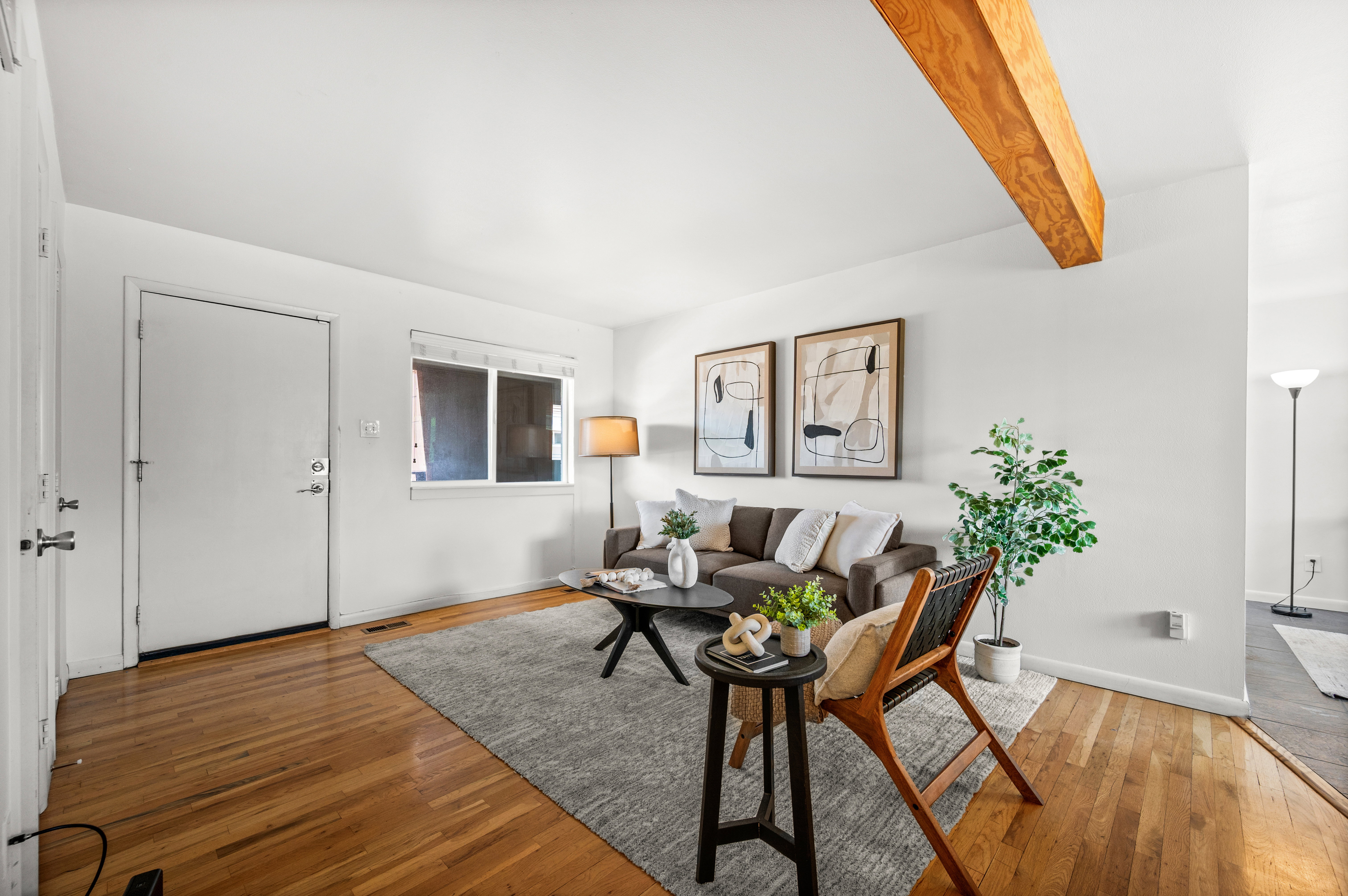 Bright living area featuring wood-finish flooring, white walls, a large window, a prominent exposed wood beam, and a neutral area rug