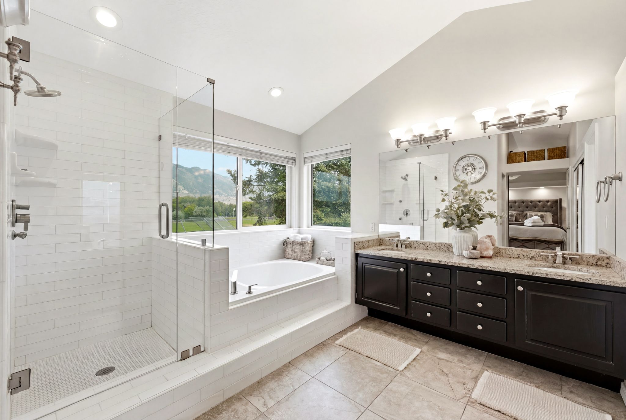 Spacious bathroom featuring a glass-enclosed shower with white subway tile, a built-in bathtub with large windows, a double vanity with dark cabinetry, and stone countertops