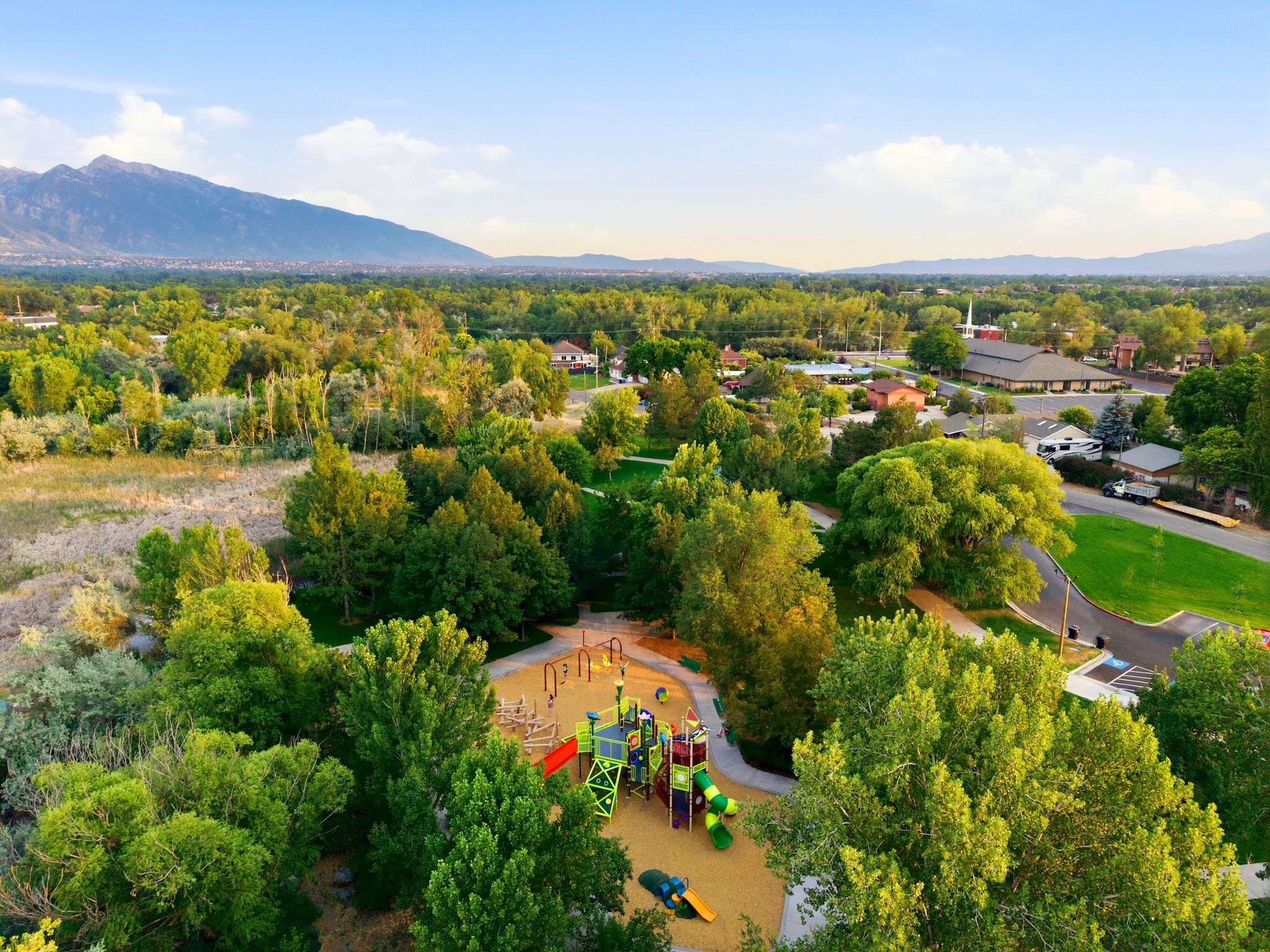 Community playground featuring a multi-level structure with slides, climbing elements, and swings, surrounded by mature trees