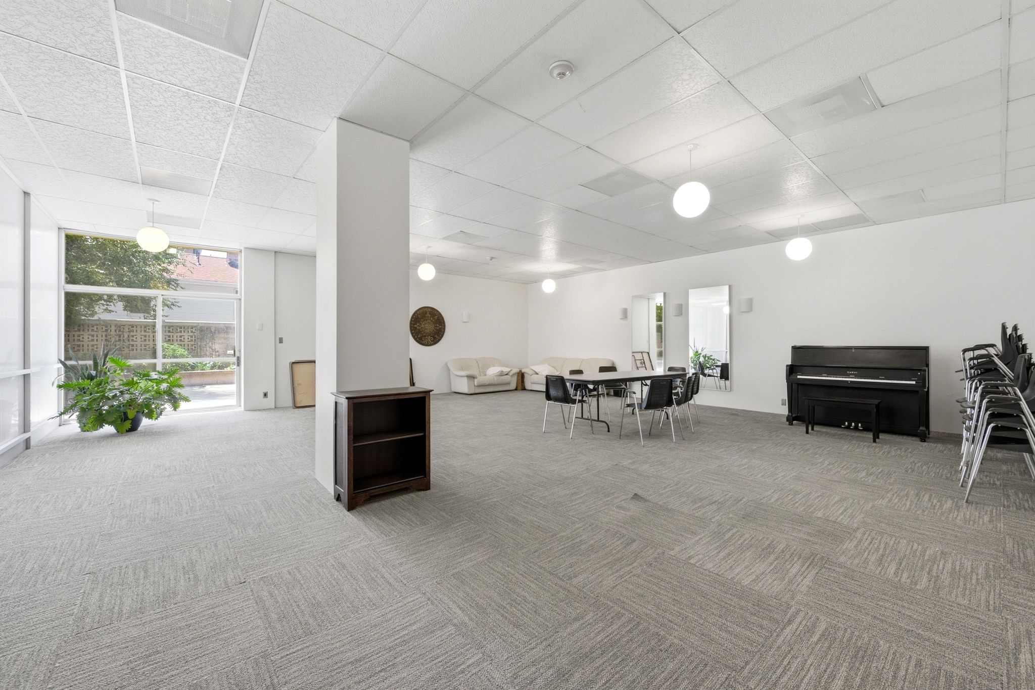 Spacious common room featuring light gray patterned carpeting, recessed ceiling lighting, and a prominent white architectural column