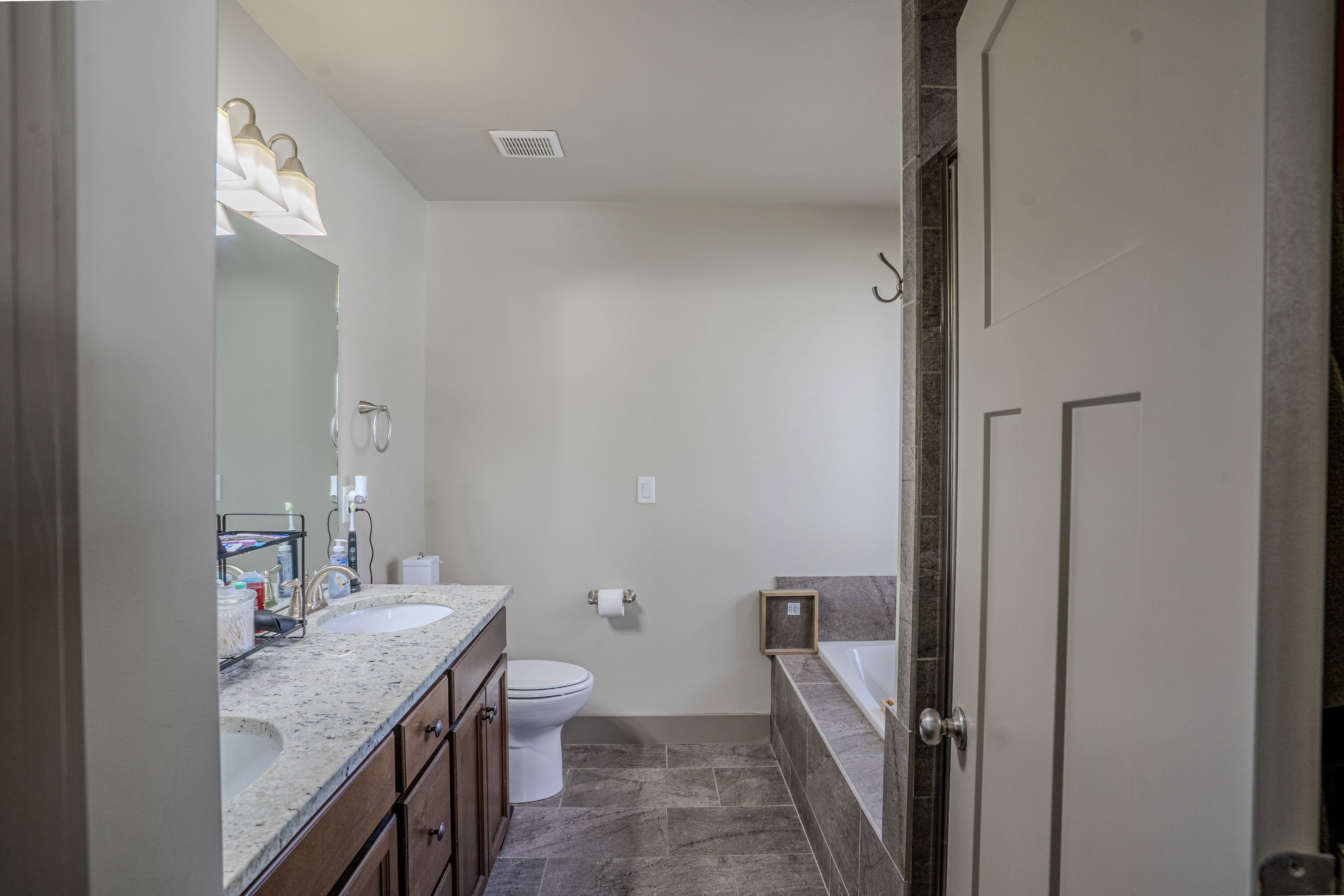 Double vanity with undermount sinks and light-toned countertop, wood-finish cabinetry, a drop-in tub with tile surround, and large format tile flooring