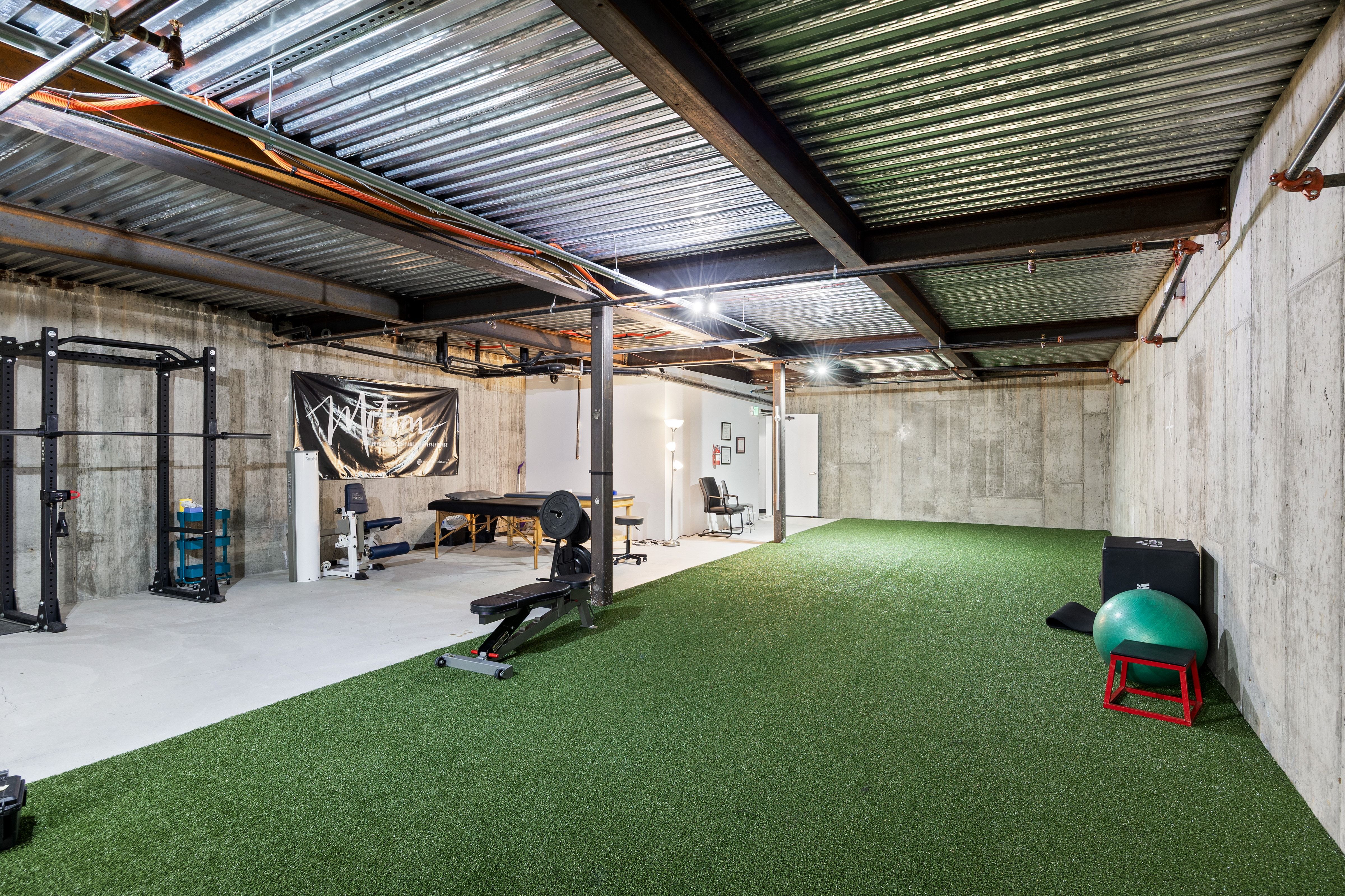 Spacious open area featuring a green turf floor section, exposed structural ceiling, concrete walls, and recessed lighting