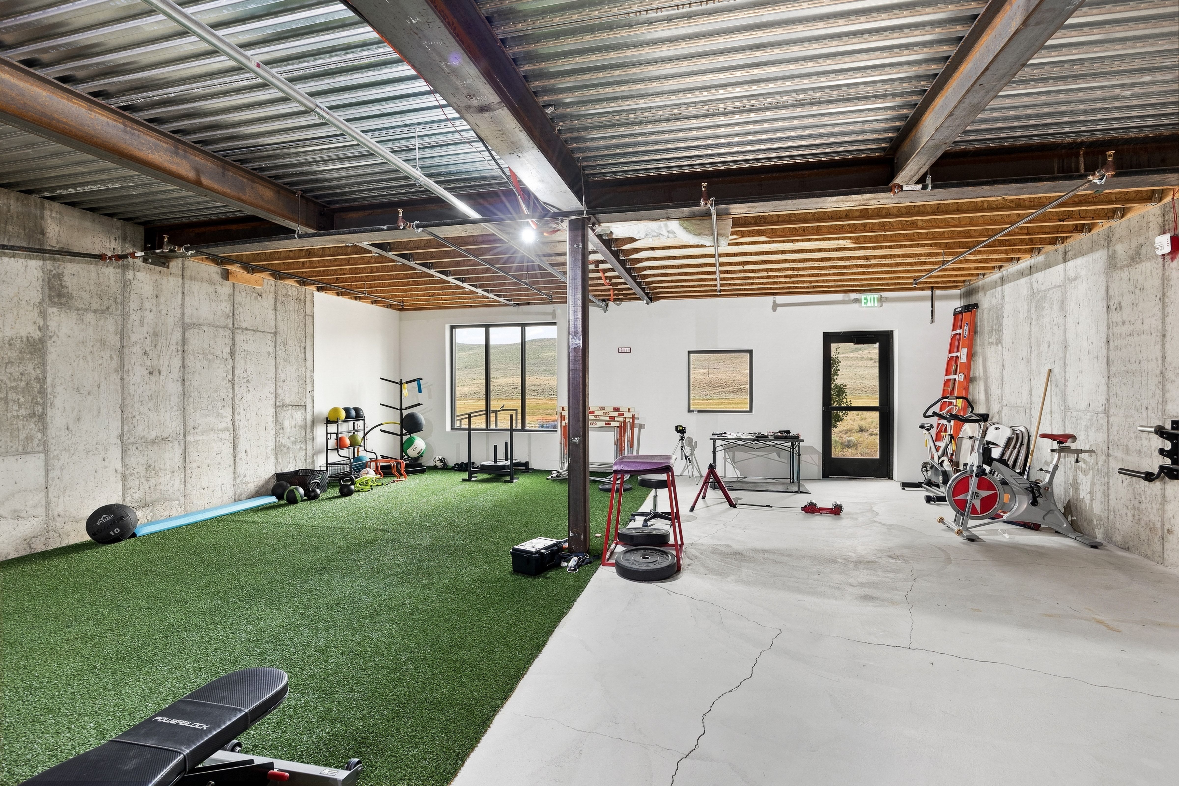 Versatile lower-level space featuring a partial concrete floor, partial green turf flooring, exposed metal ceiling joists, and concrete wall finishes