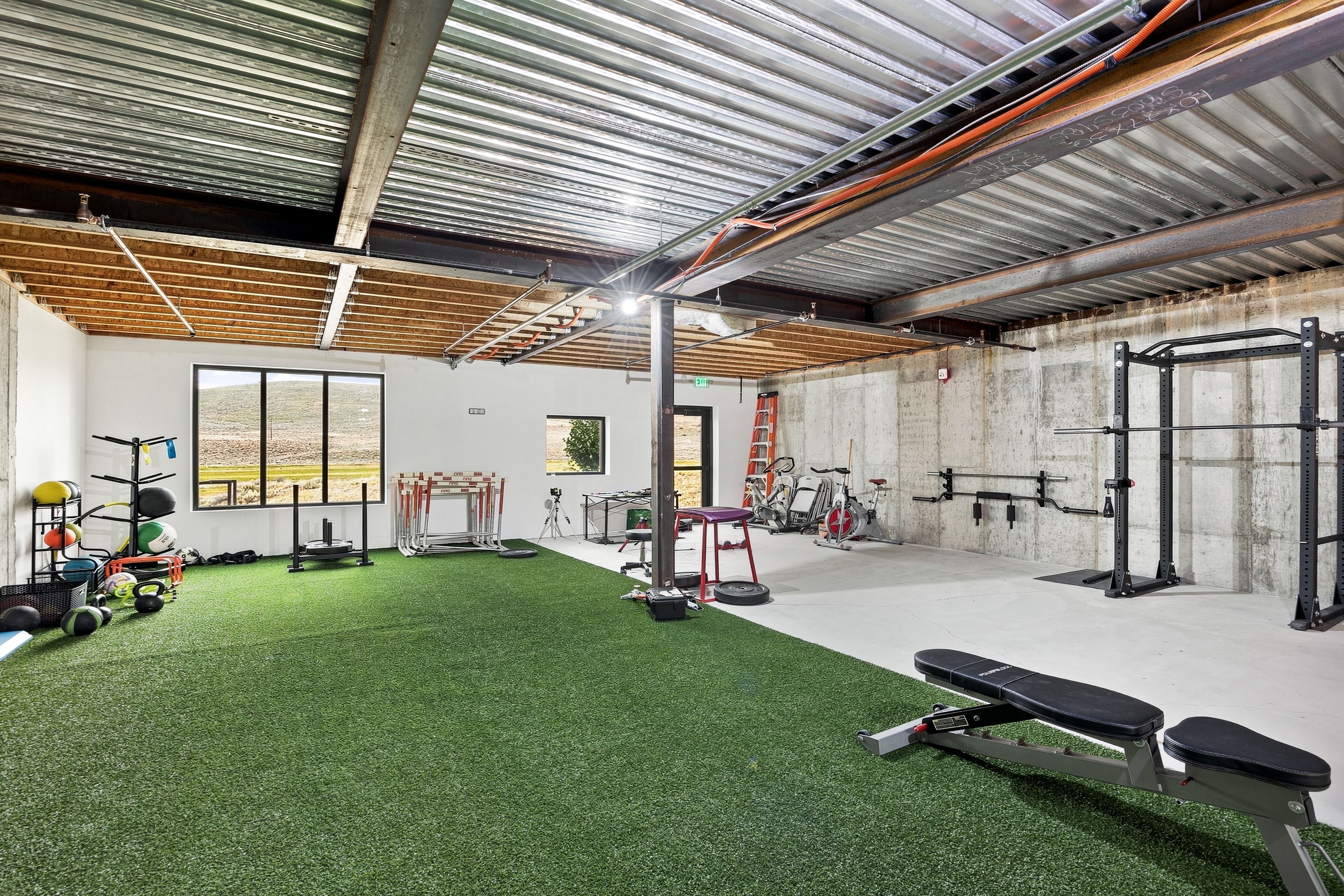 Spacious athletic facility featuring a green turf area, exposed corrugated metal ceiling, concrete walls, and large windows providing natural light