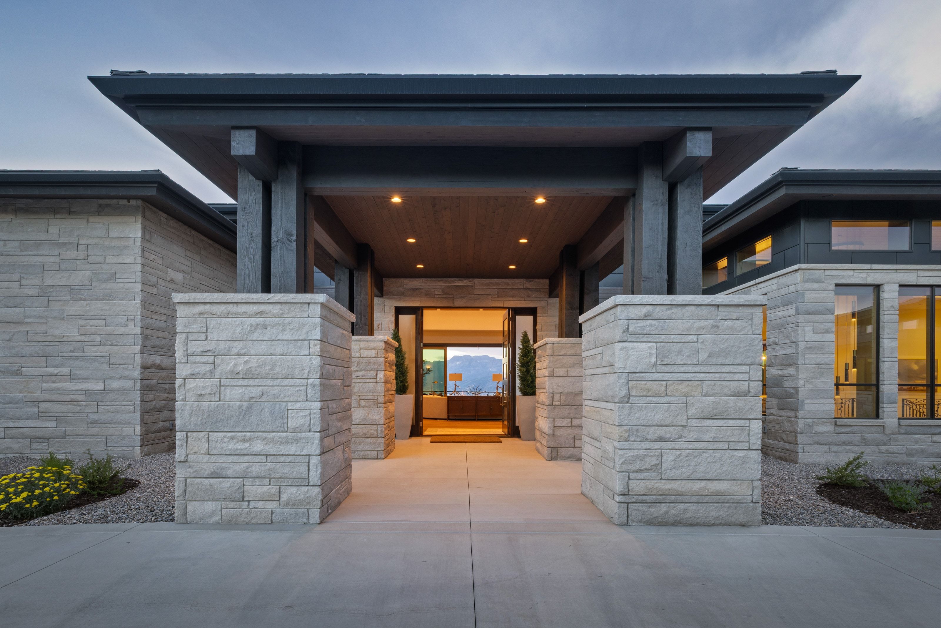 Modern architectural facade featuring stone block walls, a covered entry with recessed lighting, dark wood-finish support beams, and large windows with black frames