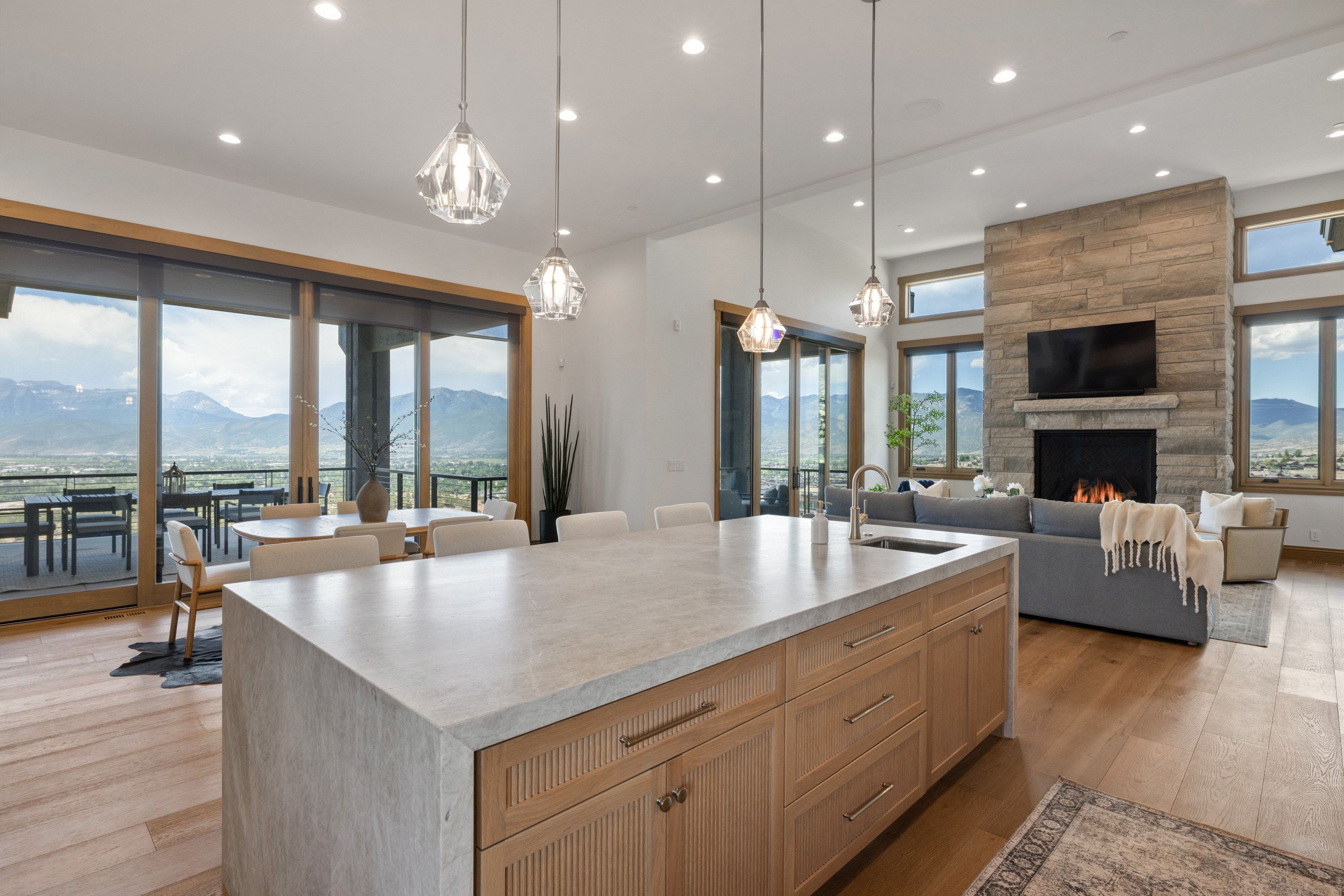 Open-concept living space featuring a large kitchen island with light-colored countertop, wood-finish flooring, a stone-clad fireplace, and expansive glass sliding doors opening to an outdoor patio with mountain scenery
