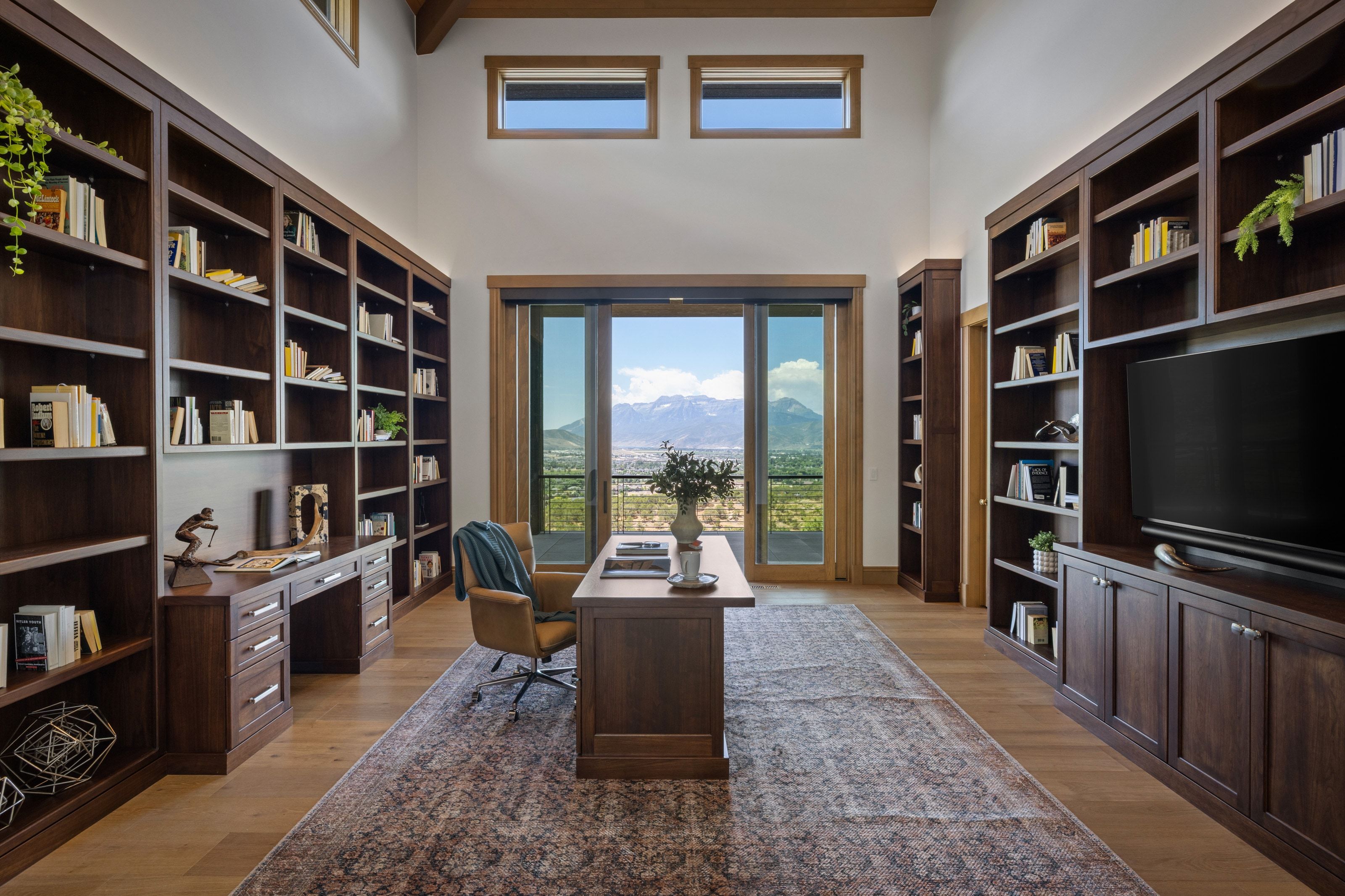 Spacious room featuring extensive built-in dark wood shelving and cabinetry, a central work desk, light wood-finish flooring, and a glass sliding door opening to a mountain and valley backdrop