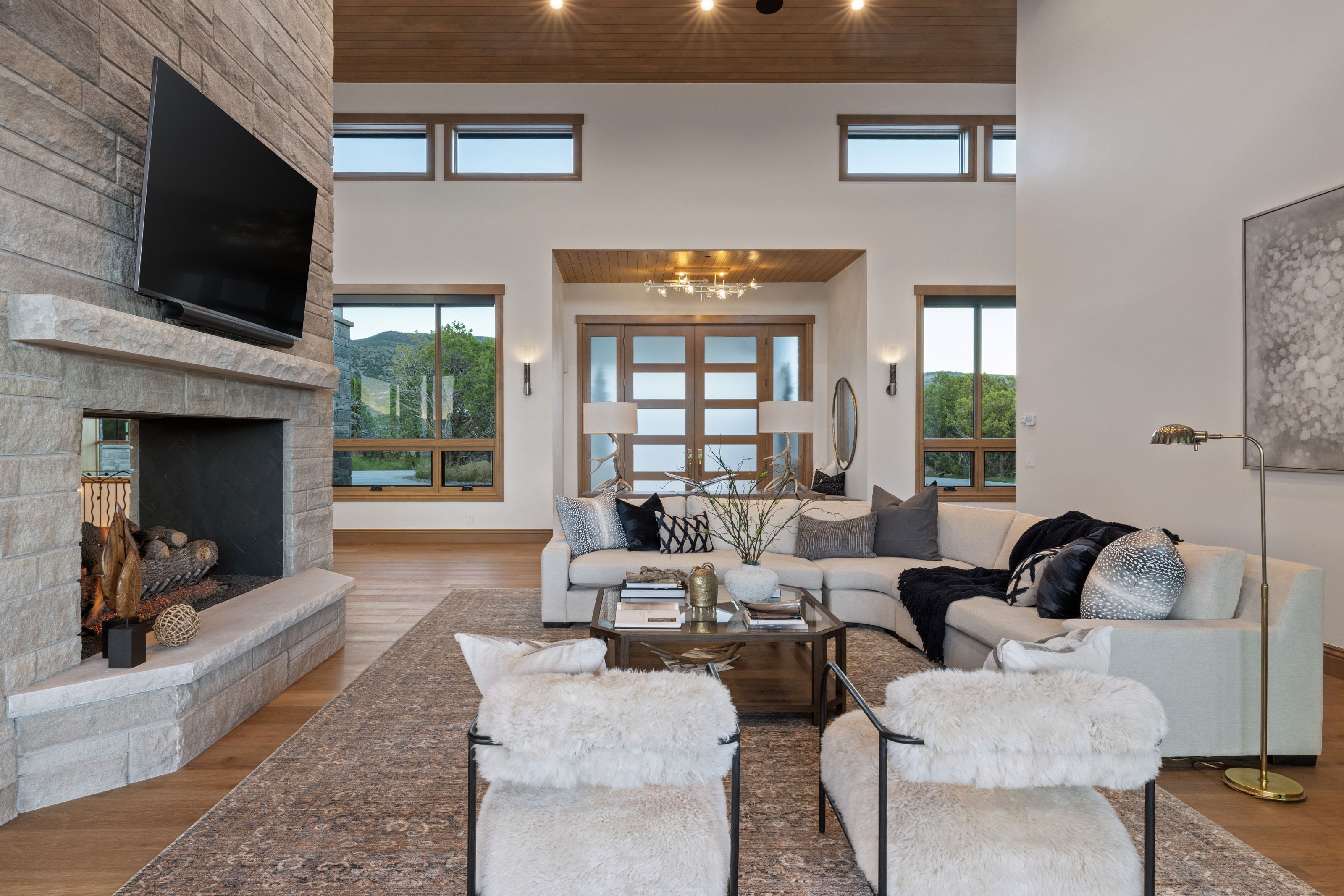 Spacious living area featuring a stacked stone fireplace, wood-finish flooring, high ceilings with recessed lighting, and multiple large windows