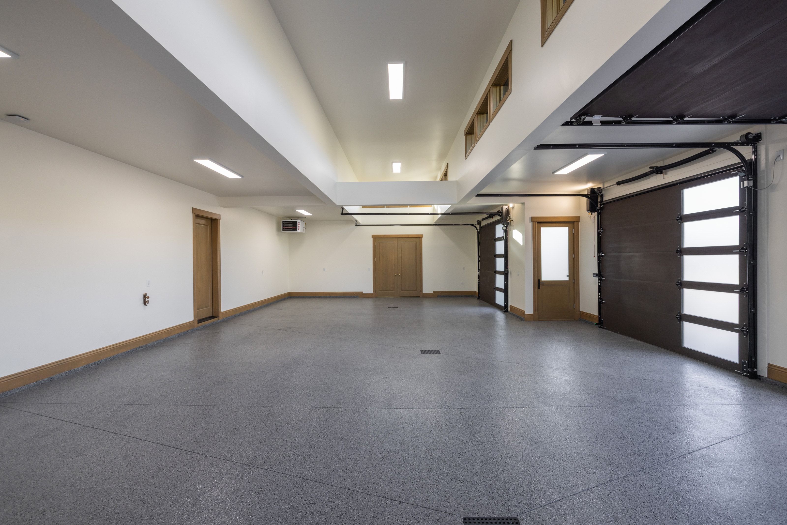 Spacious garage featuring a speckled epoxy floor, multiple overhead garage doors with frosted glass panels, warm wood-finish trim, and recessed linear lighting fixtures