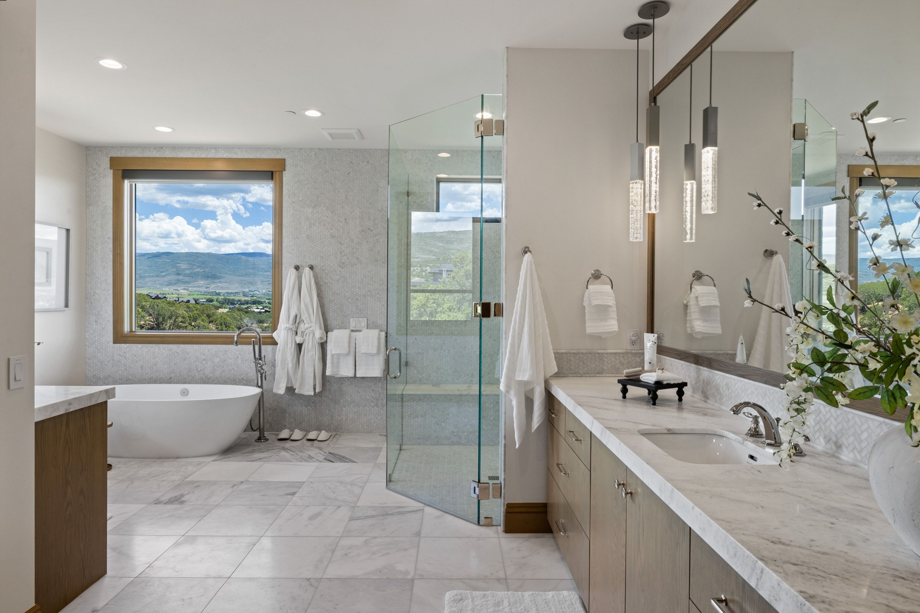 Spacious bathroom featuring a freestanding soaking tub, large window with outdoor scenery, glass-enclosed shower, dual vanity with undermount sink, and contemporary pendant lighting