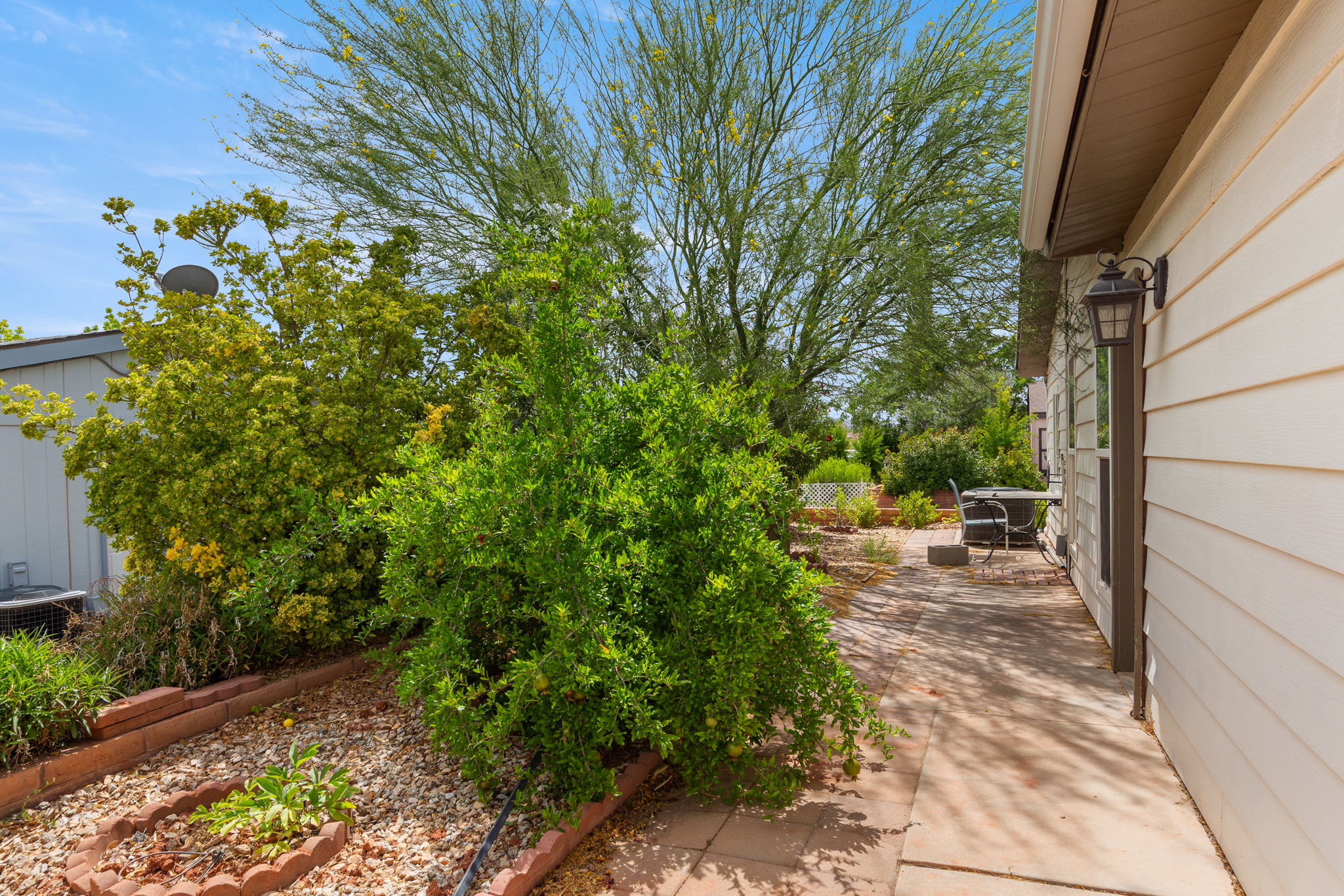 Side yard featuring mature landscaping, a paved walkway, a brick-edged planting bed, and exterior siding with an attached sconce light