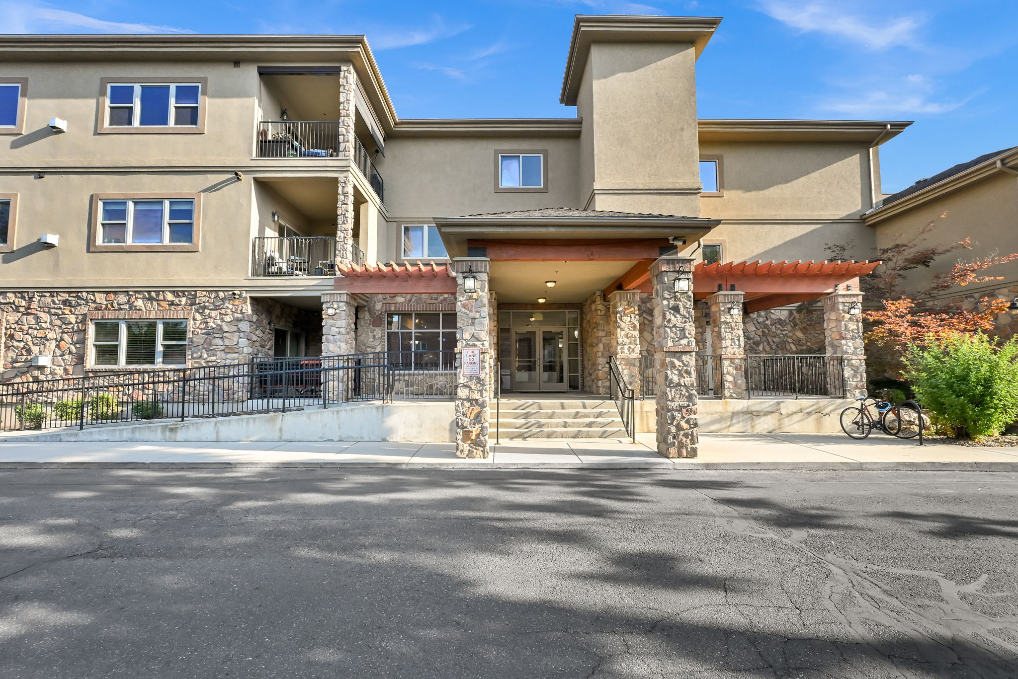 Main Entrance: Multistory building exterior featuring a prominent stone facade, multiple private balconies with metal railings, a covered entry with stone columns, and a concrete ramp with metal handrails