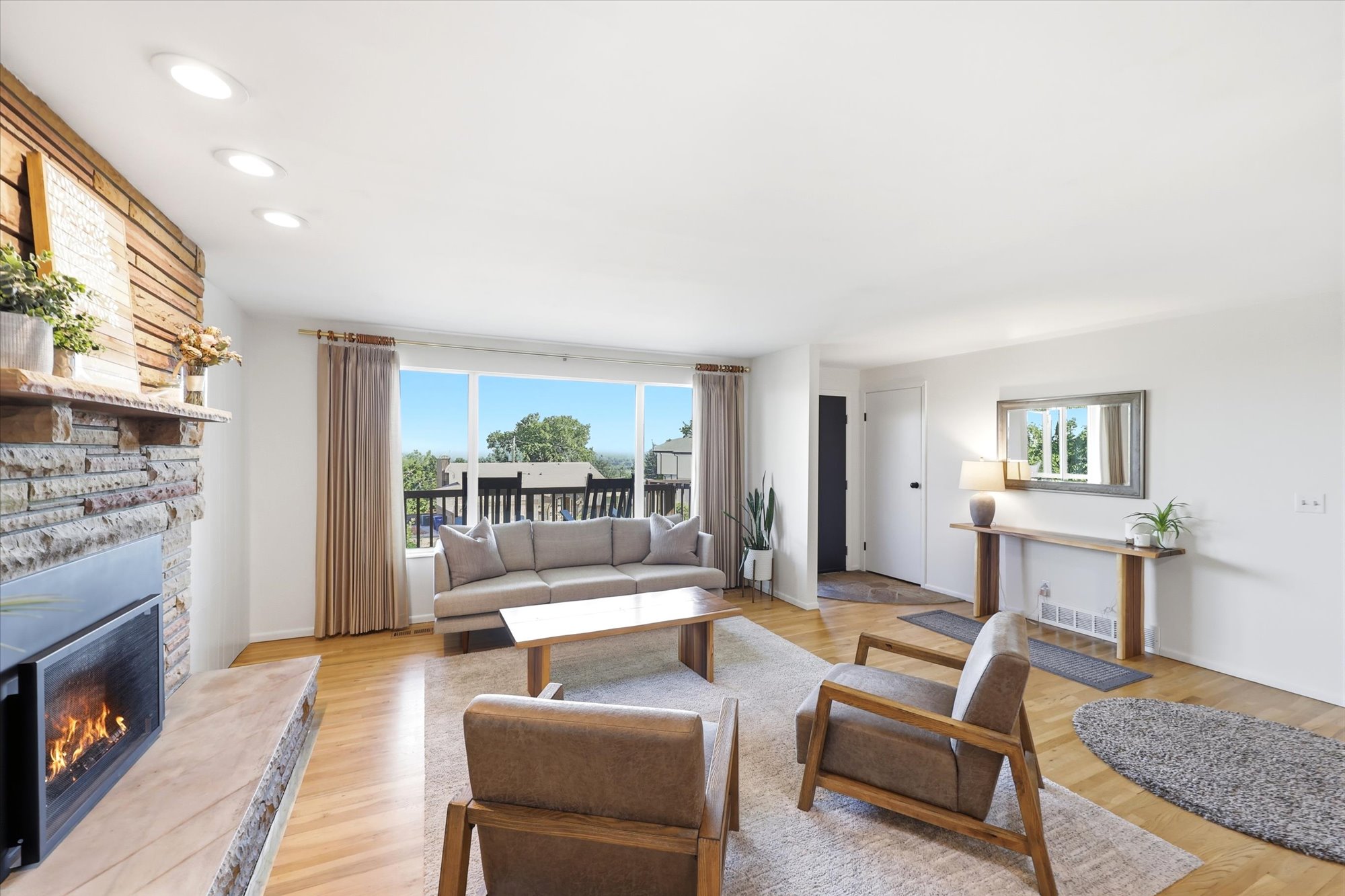 Open-concept living area featuring wood-finish flooring, a stone fireplace with a raised hearth, and a full wall of windows