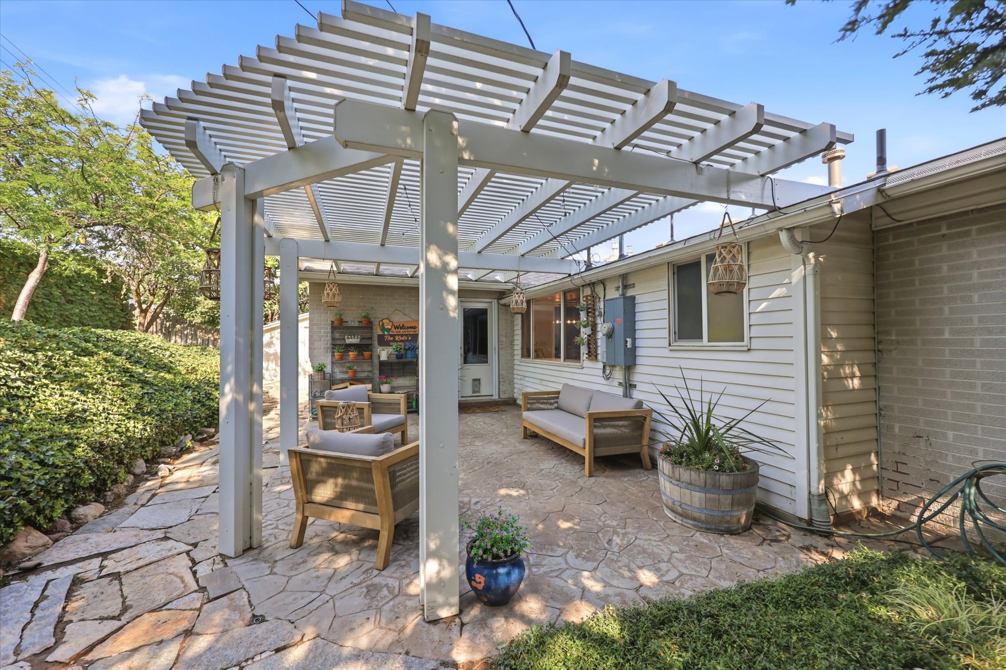 Expansive flagstone patio beneath a louvered pergola, featuring white siding, a brick exterior wall, and surrounding mature landscaping