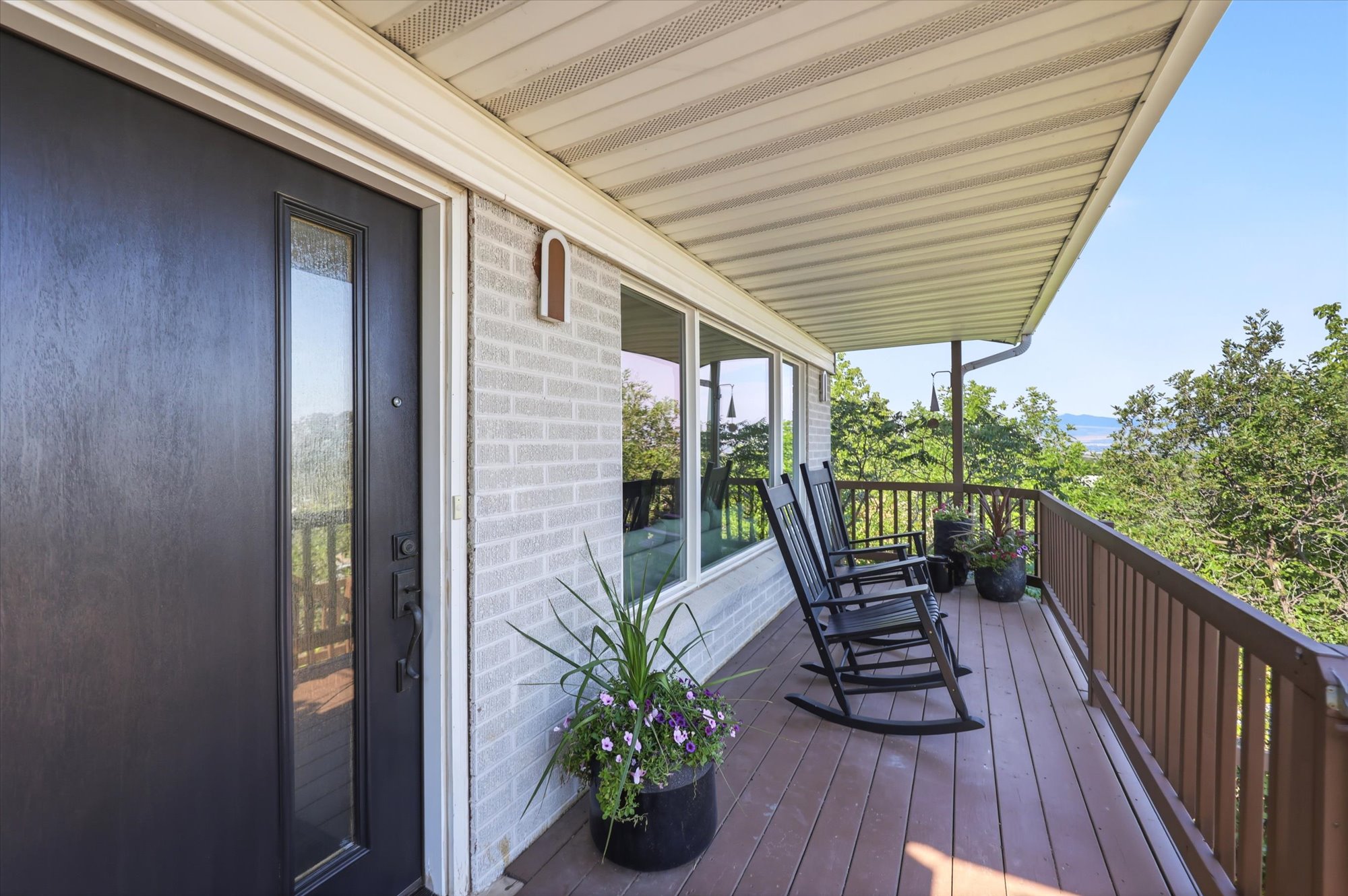 Expansive covered porch featuring wood-finish decking, a robust wood railing, a modern dark-paneled door with vertical window, a light brick exterior, and a series of large windows