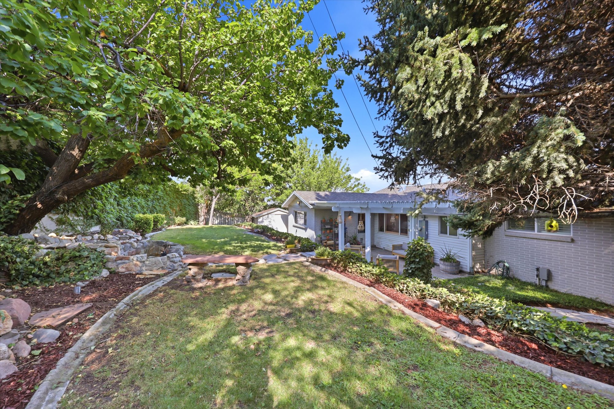 Expansive backyard featuring a manicured lawn, established shade trees, a multi-tiered rock garden, and a stone bench